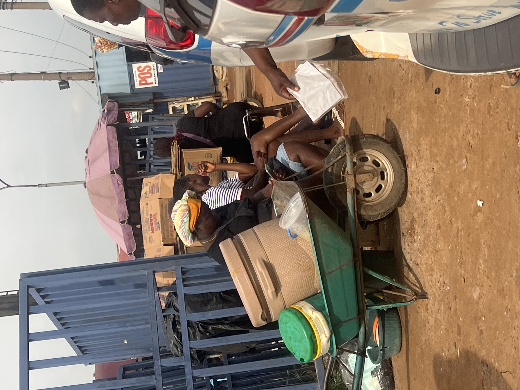 This is a medium shot, outdoor photograph taken during the day in Onitsha, Nigeria. The scene appears to be a street market or roadside commercial area.
In the foreground, a green wheelbarrow is positioned diagonally, angled from the lower left to the center right. It is filled with various containers, including a large, tan cooler or insulated container with a lid, and a smaller green plastic container with a yellow and white lid. To the left of the wheelbarrow, a dark blue metal gate or fence dominates the left side of the frame. Behind the fence, black trash bags are visible.
Several people are present in the scene. To the left, a woman wearing a colorful headwrap and an orange headscarf is seated, looking down. She is wearing a dark shirt. To her right, partially visible, is another person in a striped shirt. Further back, two women are seated on a bench under a pink and purple umbrella. One woman in a black dress is looking towards the right, and another in a striped shirt is looking towards the left.
In the background, several stalls or structures are visible. One is a blue corrugated metal structure with a sign that says "POS" and "AVAILABLE HERE." There are also stacks of cardboard boxes. A white vehicle with blue and red trim is parked on the right side of the frame, and a person's arm is visible extending from the vehicle, holding several white papers.
The ground is covered in reddish-brown dirt, typical of the region, with some scattered debris. The overall lighting suggests it is a bright, possibly overcast day.