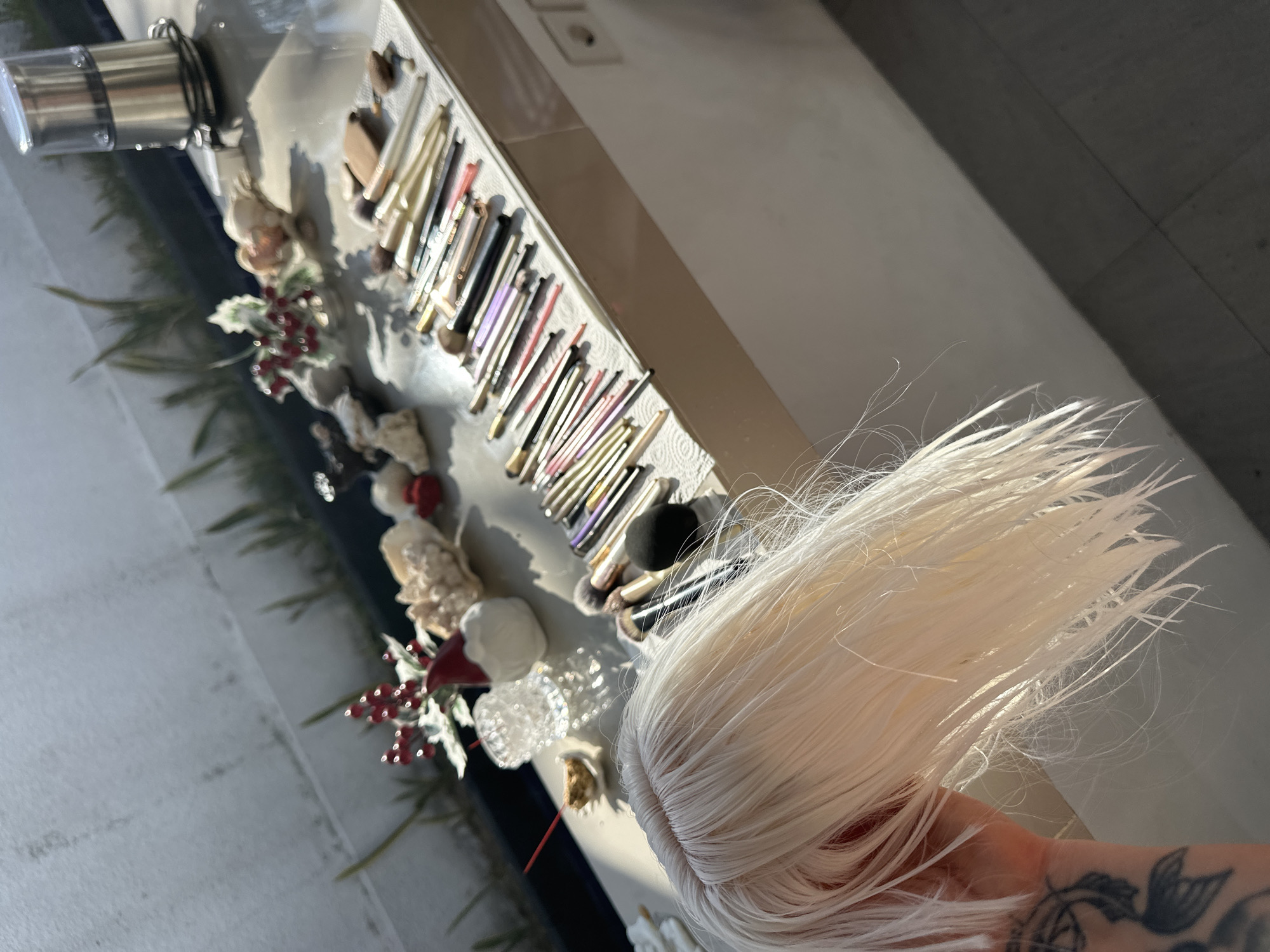 The image captures a scene on a table, possibly in an outdoor setting given the sunlight. In the foreground, a person's hand, adorned with a tattoo, is holding a portion of a pale blonde wig. The wig appears wet, with strands fanned out.
The table itself is covered with various items. A prominent collection of makeup brushes is laid out on a white paper towel. The brushes vary in size and color, with some having gold or silver handles. Adjacent to the brushes, there are decorative elements including what appear to be seashells, a small red object resembling holly berries, and a miniature gnome figurine. A crystal votive holder and an incense stick are also visible. In the background, a metallic appliance resembling a grinder sits on the table. The overall environment suggests a preparation or styling area, potentially in a residential setting in North Kuta, Indonesia, characterized by the sunlight and outdoor ambiance.