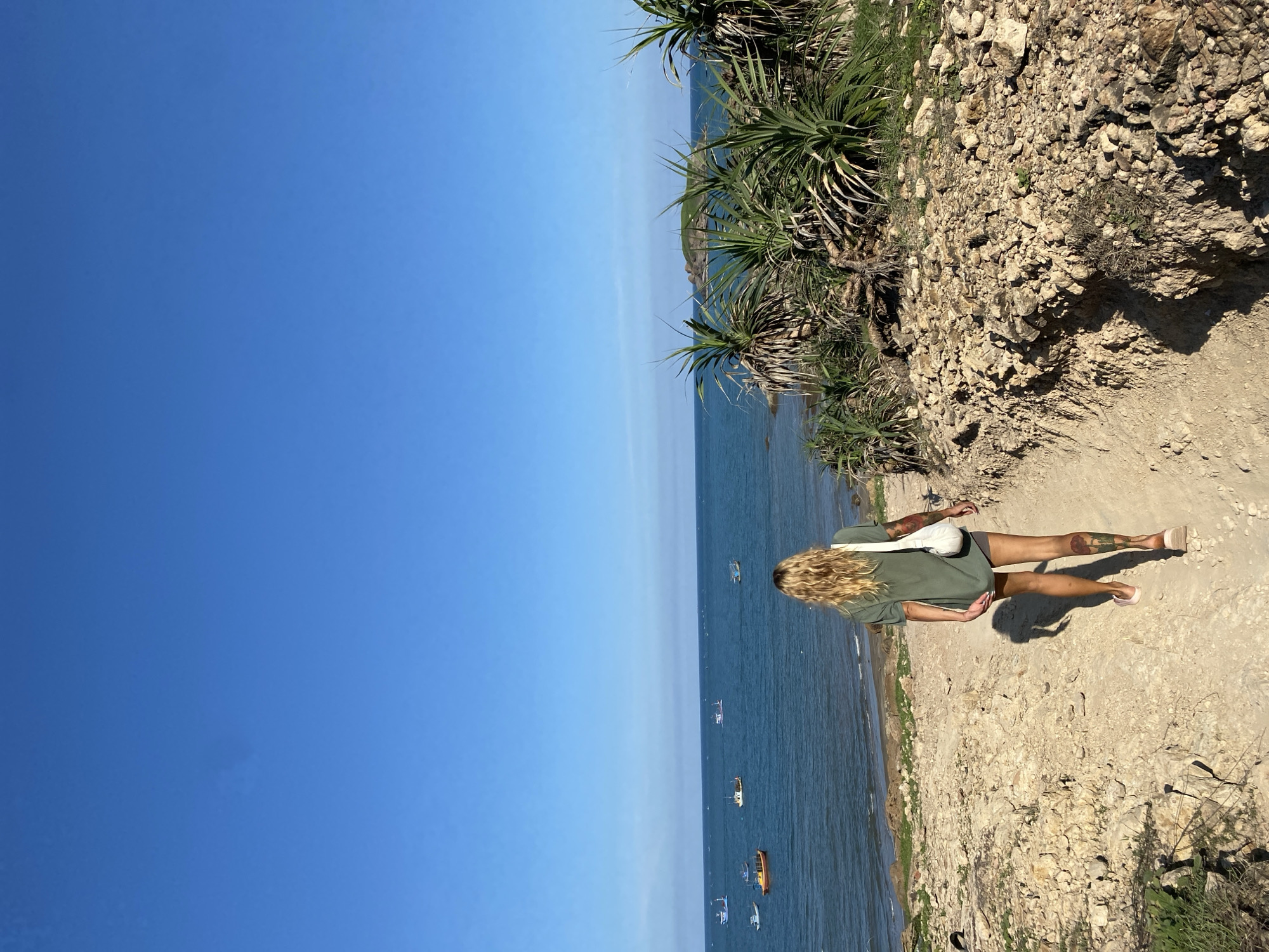 A woman with long blonde hair walks away from the camera on a sandy path leading down towards the ocean. She is wearing a green t-shirt and grey shorts, and carrying a white bag over her shoulder. Her legs are tattooed. The path is lined with rocky terrain and some sparse, spiky green plants. In the distance, the blue ocean is visible with several small fishing boats on the water. The sky is clear and blue. The scene appears to be a coastal area in Mũi Né, Vietnam.