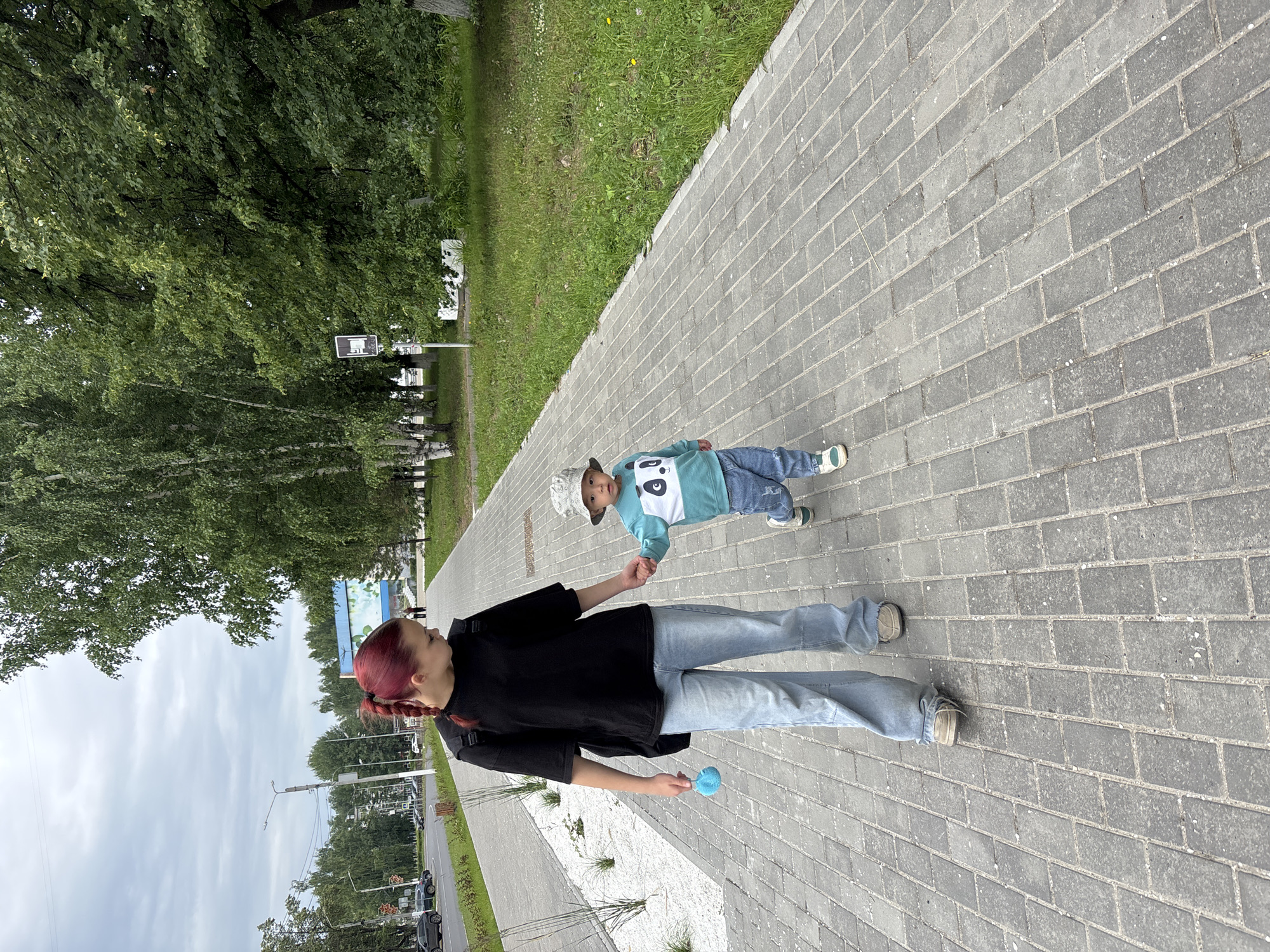 A young woman with red hair tied in a braid, wearing a black t-shirt, light blue baggy jeans, and grey shoes, walks hand-in-hand with a young child. The child, wearing a light blue sweatshirt with a panda design, blue jeans, a grey bucket hat, and white sneakers, is walking towards the viewer. The woman is holding a blue toy in her left hand. They are walking on a paved sidewalk. To the left of the sidewalk is a road with cars and trees lining the street. To the right of the sidewalk is a grassy area with trees. In the background, there is a building with a mural. The scene appears to be in a public area within the city of Chaikovsky, Russia.