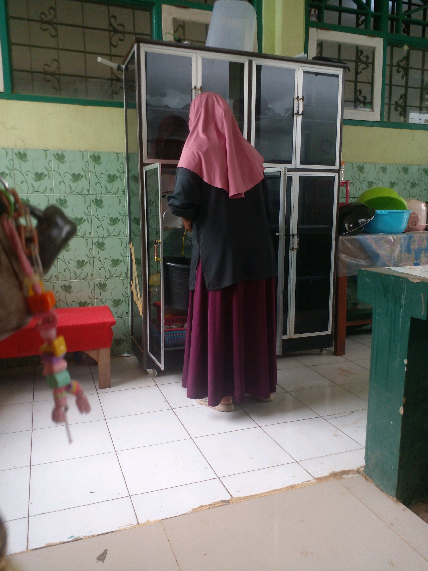 A woman wearing a pink hijab and a dark colored tunic with a long purple skirt stands facing a tall cabinet with glass doors. The cabinet is silver and black with two doors on the top half and two on the bottom half. The woman's hands are inside the cabinet. Inside the cabinet, a black bucket and blue and green bowls are visible. The cabinet sits on a white tiled floor. To the left of the cabinet, a red bench is visible. In the background, green patterned wallpaper covers the wall, and a window with green trim and a grid pattern is present. To the right of the cabinet, a green table is partially visible. The scene appears to be indoors, possibly in a storage area or kitchen.
