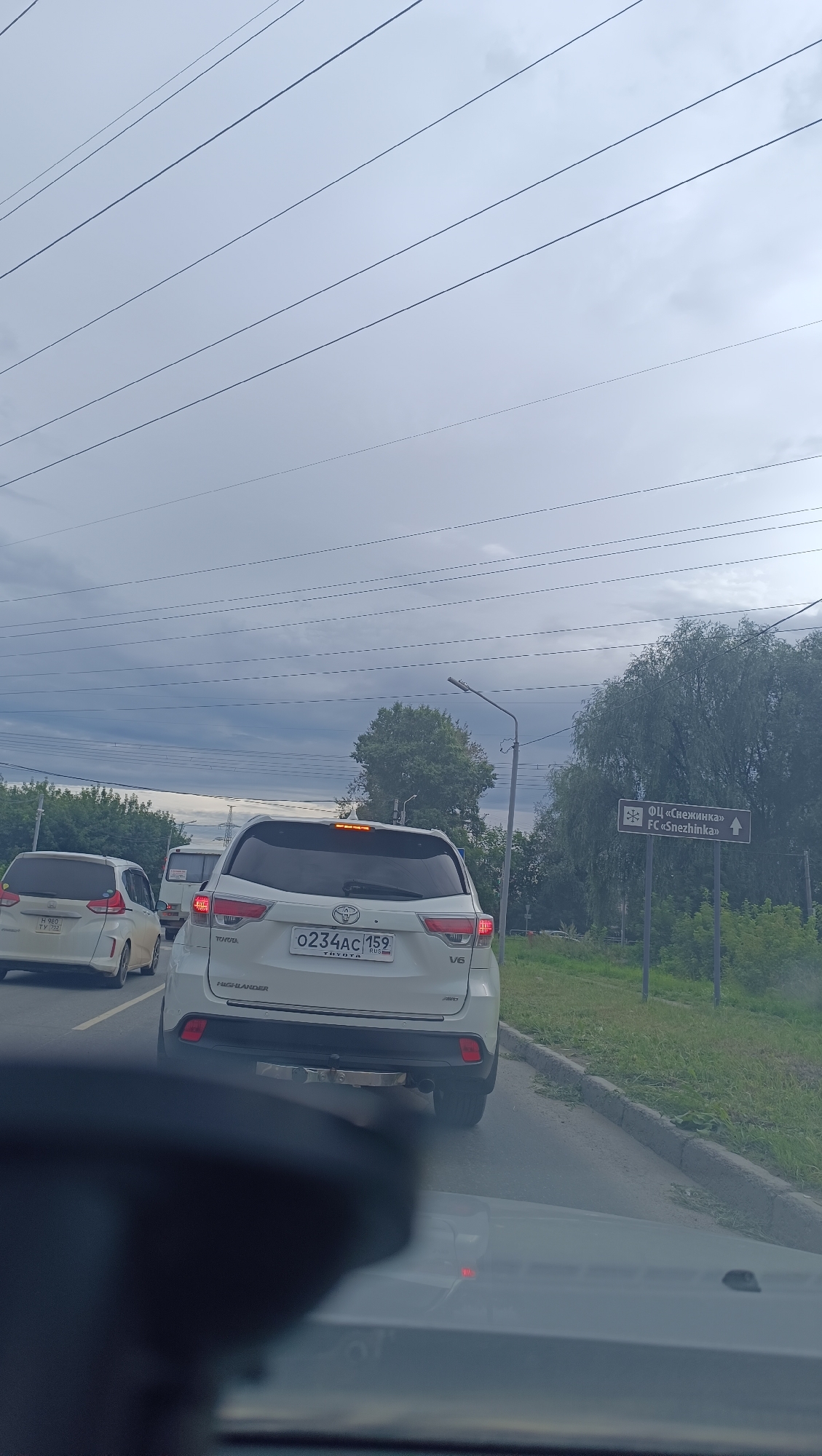 The image captures a traffic scene on a cloudy day, possibly on a road leading to or within the city of Chaikovsky, Russia. The foreground is dominated by the interior of a vehicle, with the dashboard and windshield visible. Ahead, several white cars are in traffic. The most prominent vehicle is a white Toyota Highlander with a license plate reading "0234AC 159 RUS". To its left, another white car, possibly a minivan, is visible with a license plate "H 960 TY 159". In the background, there are trees and vegetation. A road sign on the right indicates "ДЦ «Снежинка» FC «Snezhinka»" with an arrow pointing to the right, suggesting a destination or facility within the vicinity. Overhead, several power lines stretch across the sky. The overall atmosphere suggests a typical urban or suburban traffic environment.