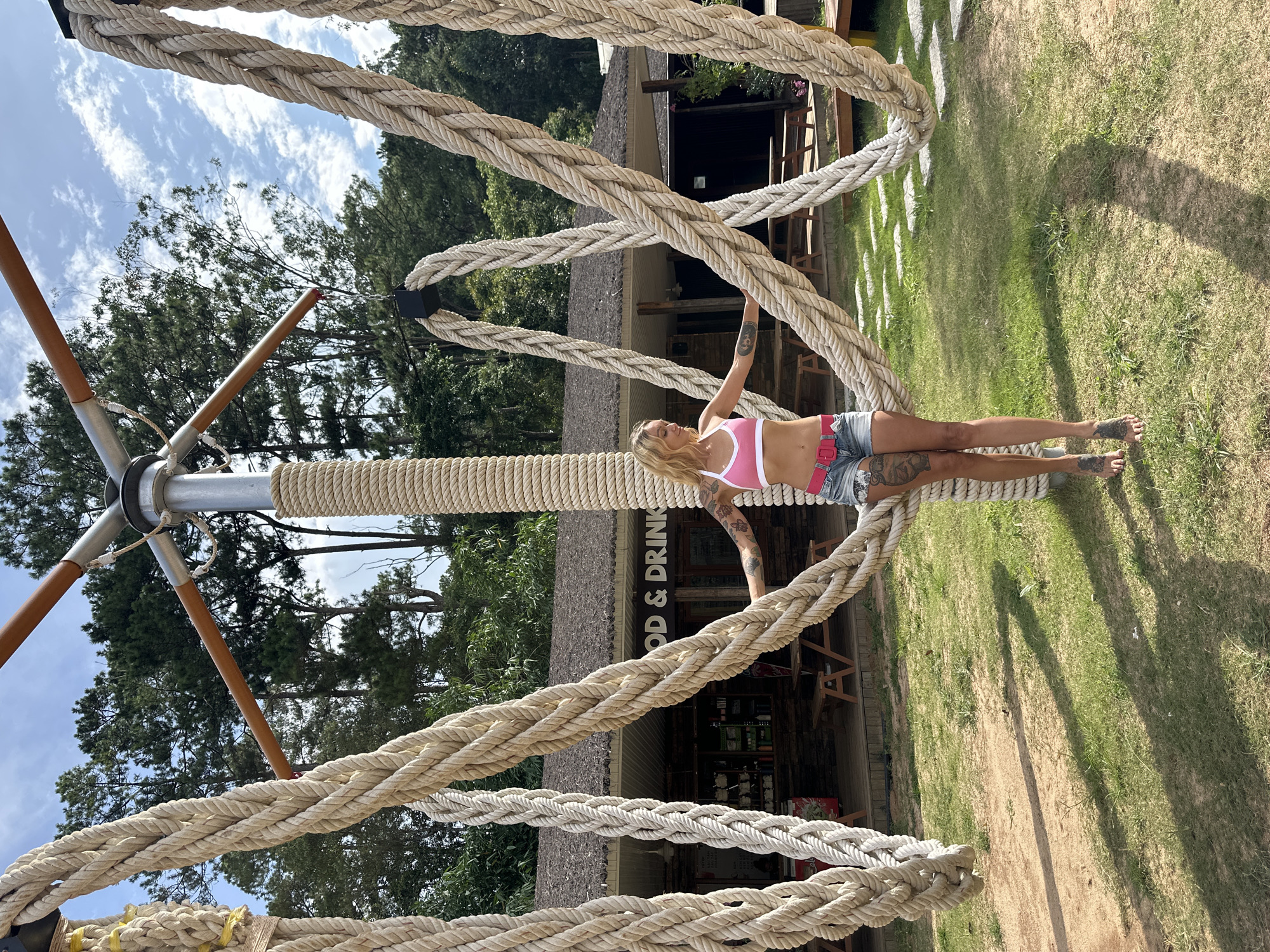 In Da Lat, Vietnam, a blonde woman with tattoos on her arms and legs stands centered in front of a structure with thick ropes. She is wearing a pink sports bra and denim shorts. The structure appears to be a large, wooden and metal frame adorned with multiple braided ropes, some of which form loops. Behind the woman is a building with a thatched roof, labeled "FOOD & DRINK," and a porch with picnic tables. The environment includes a grassy area, a dirt path, and trees in the background under a partly cloudy sky.
