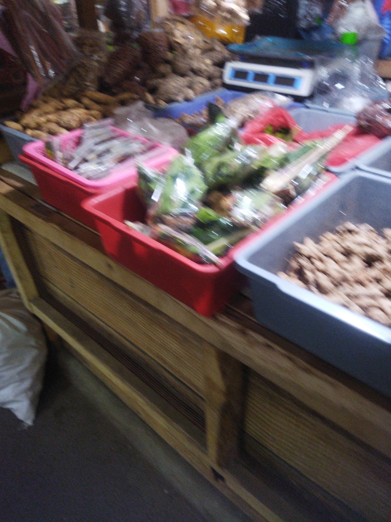 A blurred, medium shot captures a market stall in Wonogiri, Indonesia. In the foreground, a wooden counter with horizontal planks runs diagonally from the bottom left to the top right. On the counter are several containers of goods. To the left, a pink rectangular plastic basket holds items wrapped in clear plastic. To its right, a red rectangular plastic basket contains what appear to be green leafy vegetables, also wrapped in clear plastic. Further to the right, a gray rectangular plastic bin is filled with small, brown, nut-like items. In the background, out of focus, are shelves stocked with various goods, some appearing to be dried produce or spices. A digital scale with a white base and blue display is visible behind the red basket. A large, white, soft item, possibly a sack or bag, rests on the floor to the left of the counter. The lighting is dim, and the overall image has a low resolution, making fine details indistinguishable.