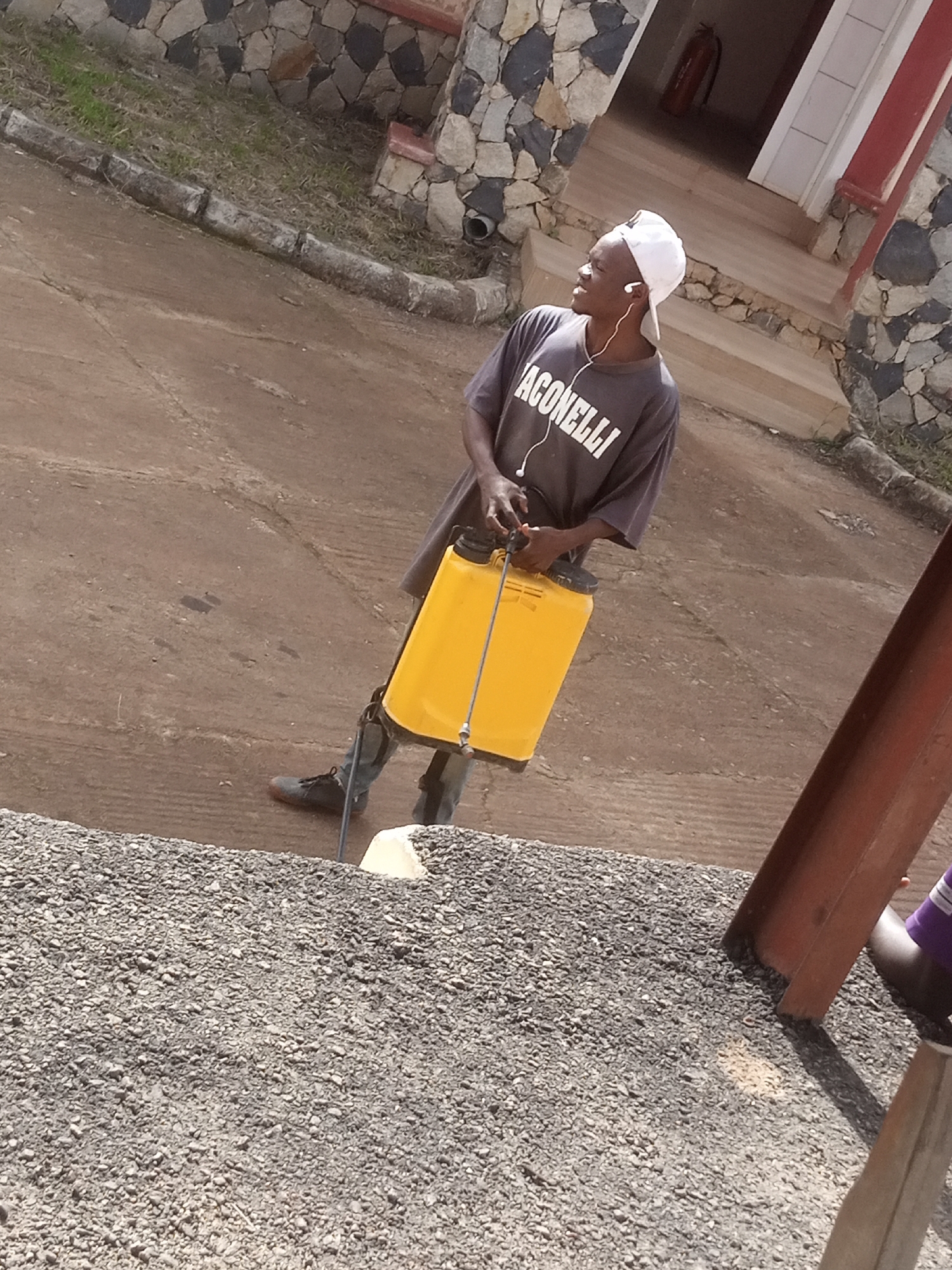 A person wearing a white baseball cap and a dark t-shirt with white lettering that reads "ACONNELLI" is standing outdoors, holding a yellow sprayer. The person is also wearing dark pants and boots. The sprayer is positioned on their right side, and they are looking to their left. The background shows a building with a stone facade and an entrance with stairs. The ground is a dirt or gravel surface. The image is taken from a low angle.