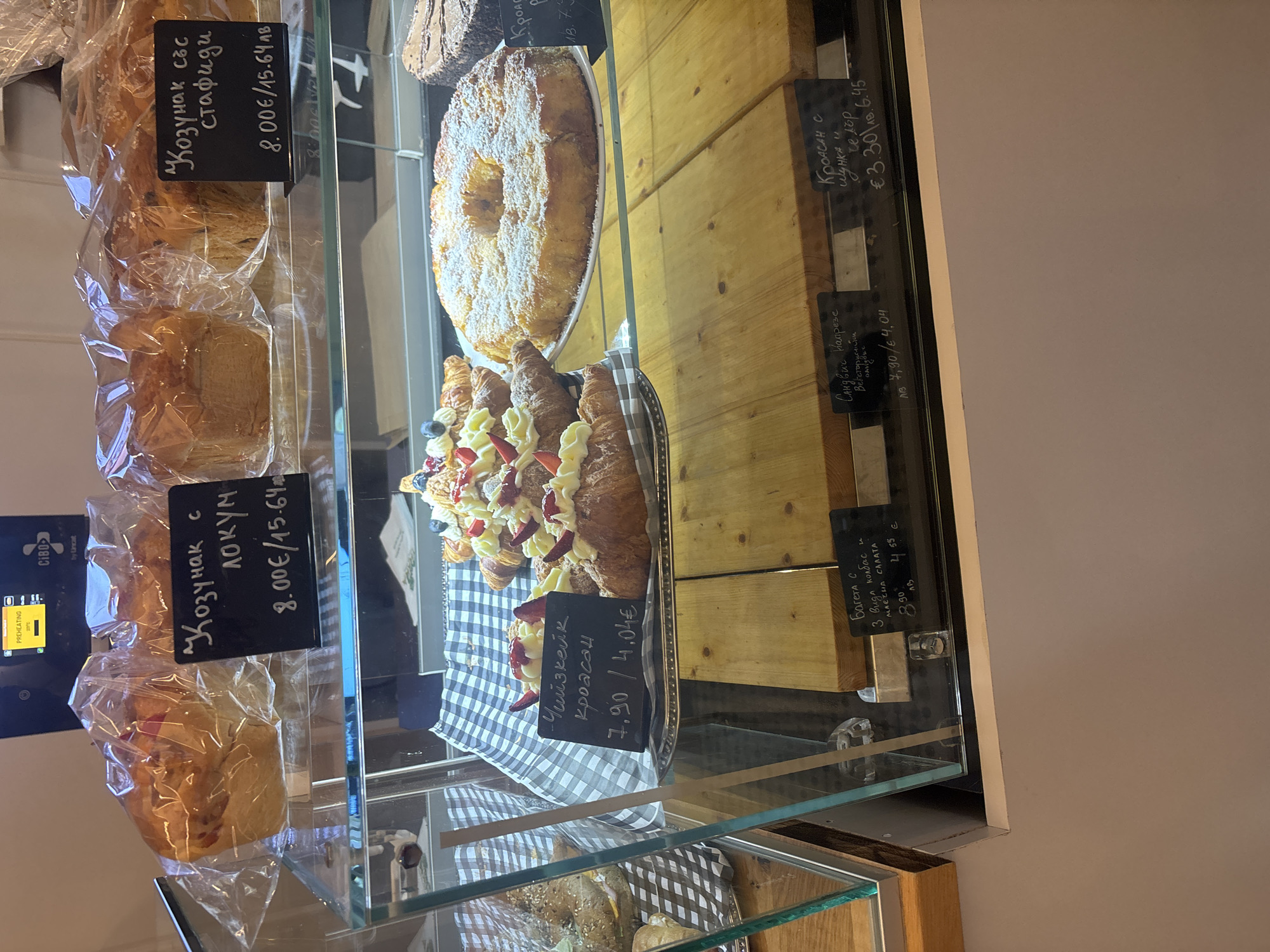 A bakery display case contains various pastries, including kozunak (a sweet bread), croissants, a cheesecake croissant, and a ring cake. The kozunak is priced at 8.00€ or 15.64лв, and the cheesecake croissant is 7,90/4.04€. A sign indicates that the baguettes with three types of sausage and milk salad cost 8.90лв or 4.55€. The pastries are displayed on a checkered cloth and wooden shelves within a glass display case. A CIBO by Lancat oven is also visible. Based on the currency (лв) used, it is likely in Burgas, Bulgaria.