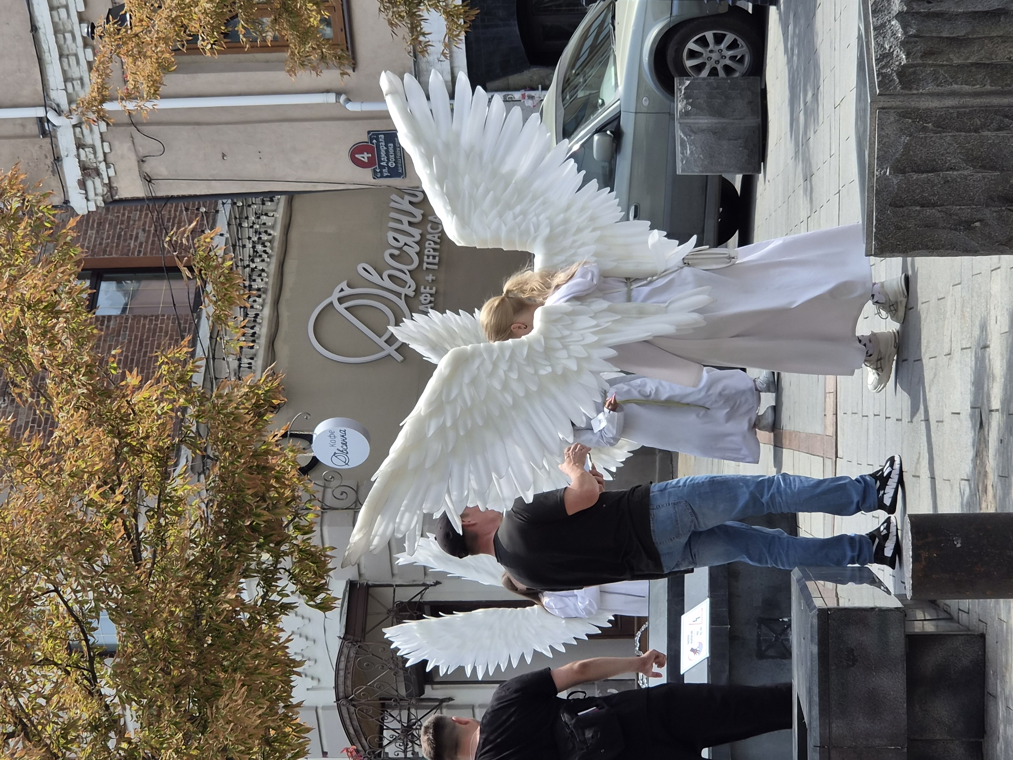 In Vladivostok, Russia, on a sunny day, two individuals are dressed as angels with large white feathered wings. One angel, facing away from the camera, wears a white robe and sneakers. The other angel, whose face is not visible, also wears a white robe and sneakers. They are positioned in a public outdoor area with paving stones underfoot. In the background, a car is parked, and a building with a sign that appears to read "Кафе Овсянка" (Cafe Ovsyanka) is visible. Trees with autumnal foliage are also present. Two men are standing to the left of the angels. One man, facing away, wears a black t-shirt and shorts, and has a backpack. The other man, also facing away, wears a black t-shirt and blue jeans. They appear to be observing the angels. The environment suggests an urban street or plaza.