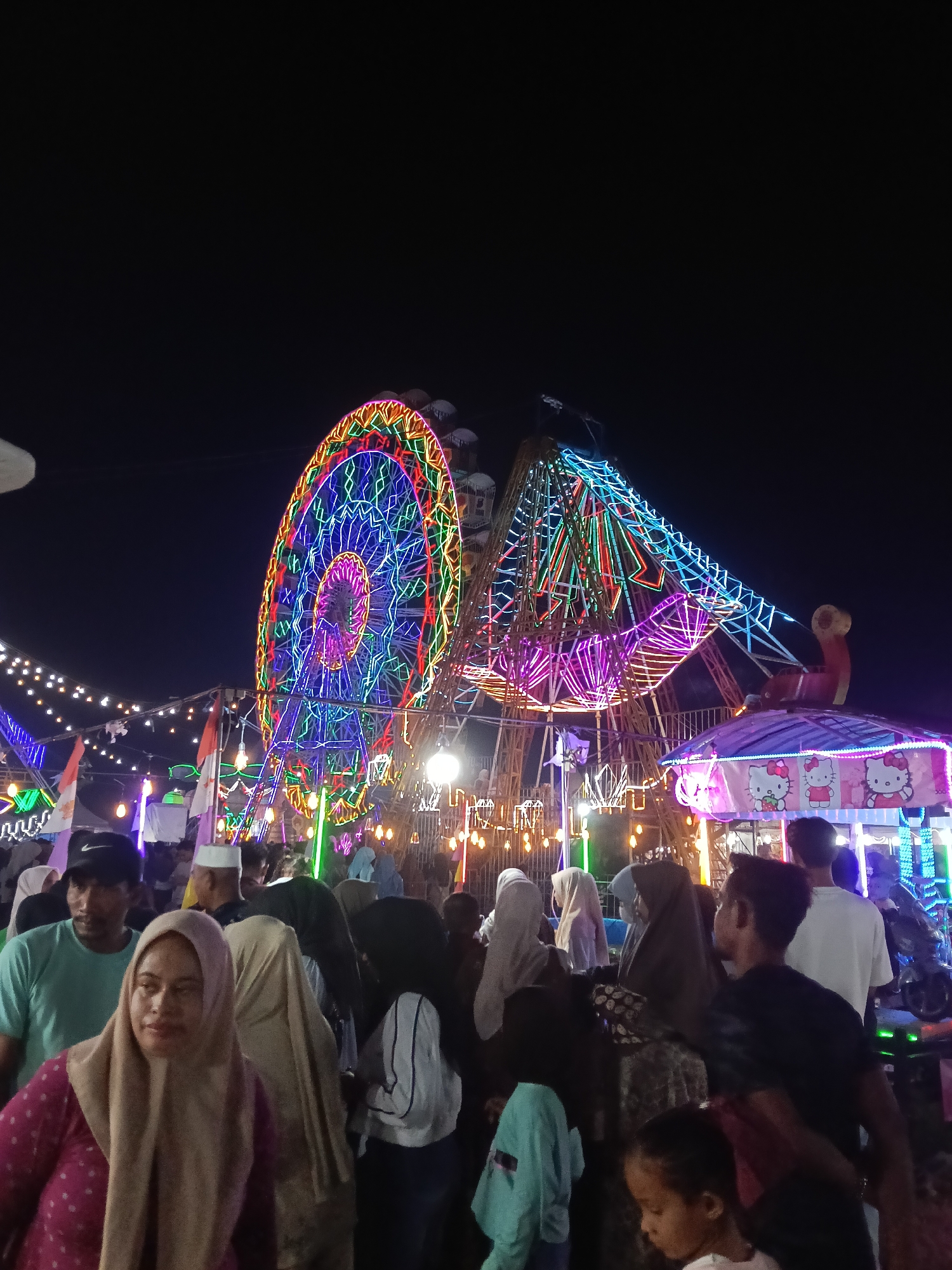 This image captures a lively scene at night in Keudepanteubreuh, Indonesia, likely during a local festival or fair. The focal point is a brightly lit Ferris wheel, adorned with a vibrant array of colorful neon lights in blue, green, red, and pink, forming intricate patterns. Adjacent to it is another amusement ride, possibly a swing ride, also illuminated with colorful lights. A smaller structure with a canopy featuring cartoon characters, specifically Hello Kitty, is visible in the foreground.
A crowd of people, dressed in casual attire and many wearing headscarves, are milling around the attractions. Some are looking towards the rides, while others appear to be walking or conversing. The atmosphere is festive and bustling, with the dark night sky serving as a backdrop to the illuminated attractions and the silhouettes of the people. The ground is dimly lit, but the lights from the rides create a colorful and somewhat diffused illumination. The presence of Indonesian flags indicates a local celebration.