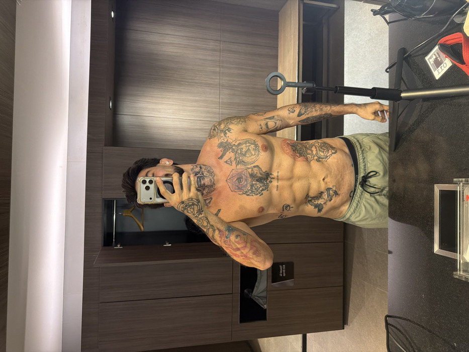 An adult male is depicted in a mirror selfie within an indoor changing room or locker facility located in Bangkok, Thailand. The man, shirtless and wearing light-colored drawstring shorts over dark underwear, stands directly facing the mirror. His torso, arms, and neck are extensively covered with blackwork tattoos featuring various designs, including geometric shapes, eyes, animals, and mythological figures, with some faint red accents. He has dark hair and a beard, with his face partially obscured by the silver-colored smartphone with a clear case that he holds in his right hand, pointing towards the mirror to capture his reflection.
The background, visible in the mirror's reflection, consists of dark wood-paneled walls or cabinetry. On the left, a cabinet door is ajar, revealing a wooden hanger inside. Below this, a recessed compartment holds a grey fabric item, and a small white sign, potentially bearing Thai script, is affixed to the inner wall. To the right, a long, light-colored wooden bench is positioned against a similar wood-paneled wall. Overhead, a continuous strip of white LED lighting illuminates the space.
In the foreground, on a dark, reflective countertop in front of the man, several objects are present. To the right, a black extendable tripod or stand with a circular top attachment is prominently displayed. Beside it are a red fabric item, possibly a bag strap or clothing, and a white rectangular card with "20L" printed on it along with a small logo. To the left, a clear rectangular container with internal contents is also visible on the countertop.