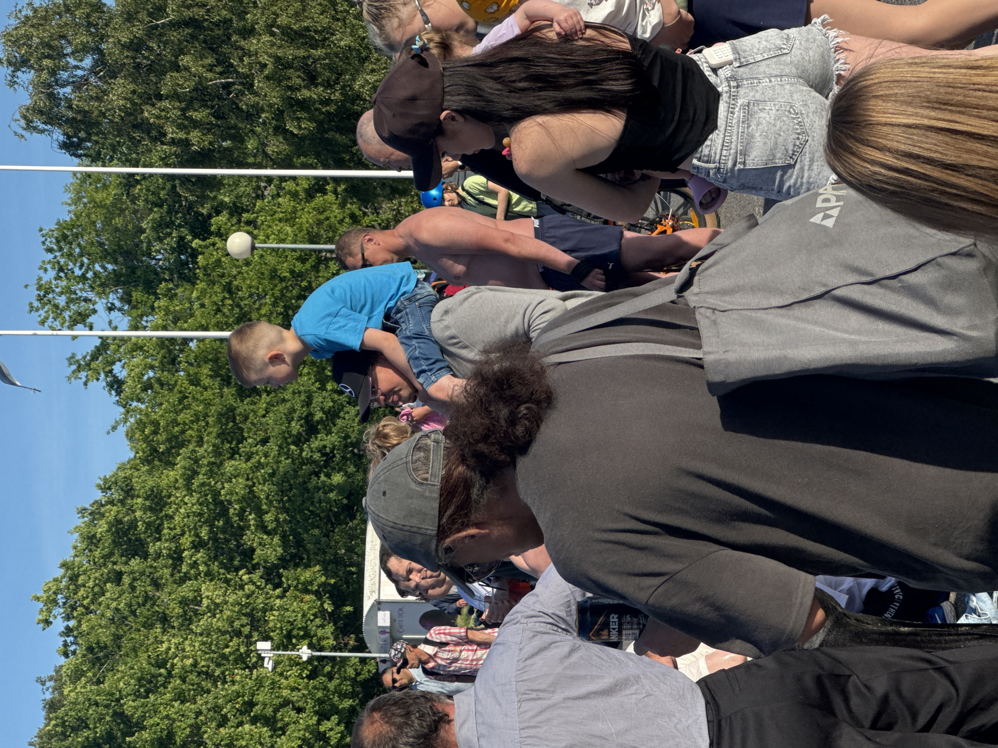 The image depicts a crowd of people outdoors, likely at an event or festival, given the presence of food stands and a festive atmosphere. The background features lush green trees and a clear blue sky. In the foreground, a young boy is seated on a man's shoulders, both dressed casually. To their left, a person in a grey shirt and black pants is visible, holding a can of "INKER" brand beverage. Another individual in the mid-ground is wearing a black baseball cap and a dark tank top, while a person next to them is holding a child. Several other individuals are scattered throughout the frame, engaged in conversation or observing their surroundings. The overall scene suggests a warm, sunny day and a relaxed, social gathering. While the specific location within Tallinn, Estonia, cannot be determined from the image alone, the context implies an outdoor public space conducive to such an event.