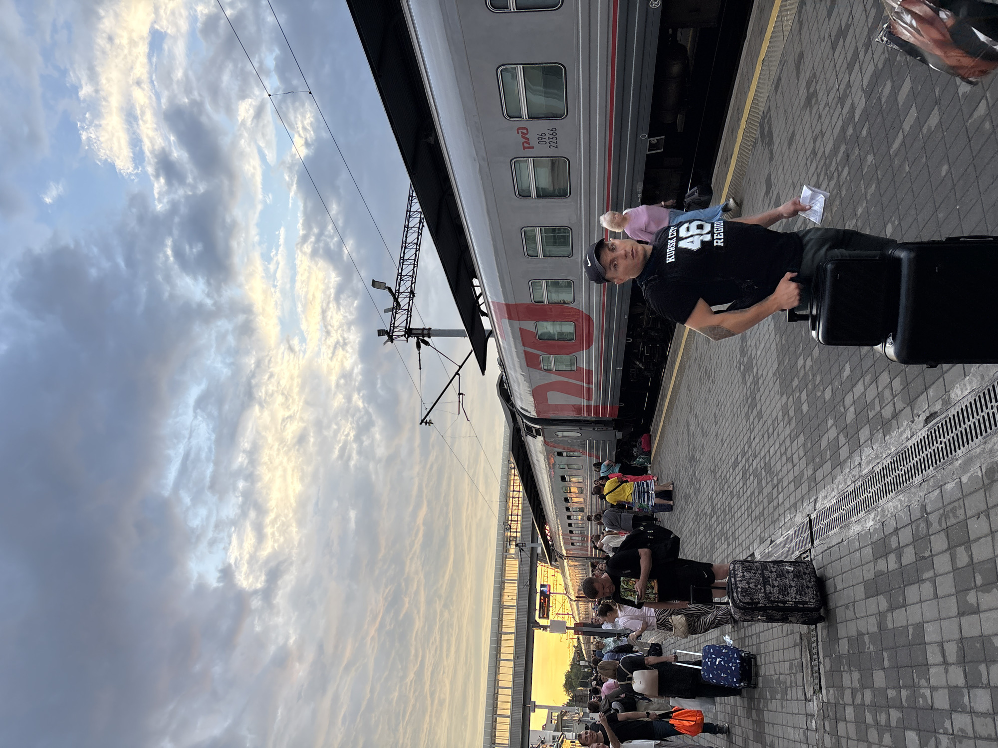 The image depicts a train station platform in Khabarovsk, Russia. A train, with "РЖД" (Russian Railways) branding, is parked alongside the platform. Several people are visible, some with luggage, suggesting they are either arriving or departing. The sky is partly cloudy, with indications of sunset or sunrise. The platform is paved with interlocking bricks, and a drain runs along its edge. In the foreground, a man wearing a black t-shirt with "KURSK CITY 46 REGION" printed on it is holding a piece of paper and standing next to two black suitcases. Other individuals are spread across the platform, some walking, others standing. A covered walkway or bridge is visible in the background, spanning across the tracks.