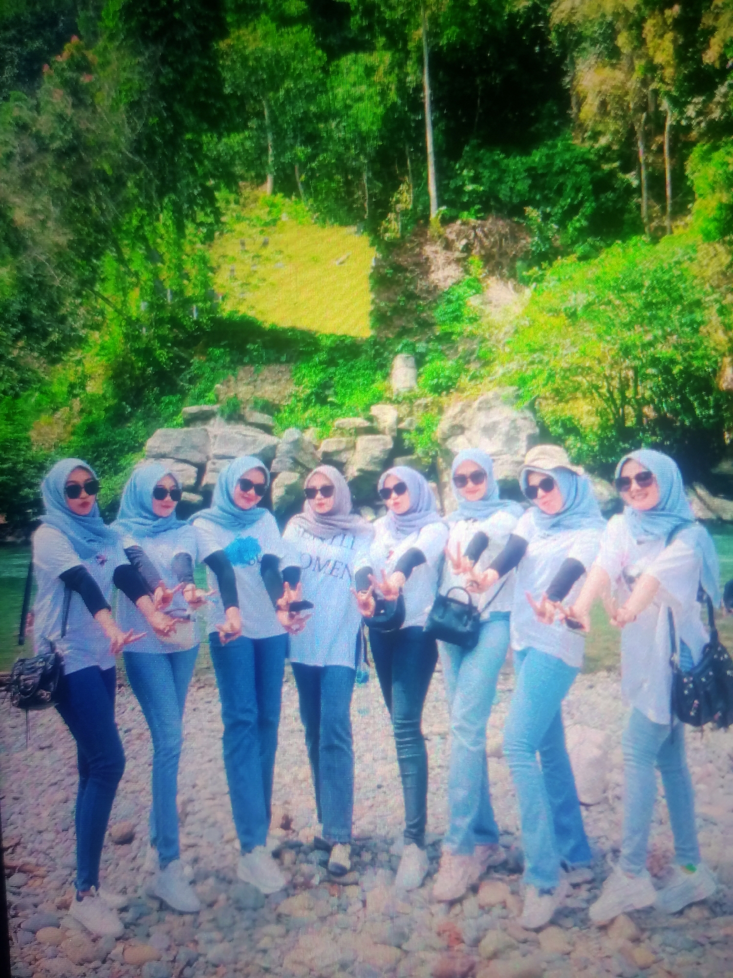 Eight women wearing light blue hijabs and sunglasses pose for a photo in front of a natural backdrop. They are all dressed in white t-shirts and blue jeans, with some wearing white sneakers and others pink sneakers. The woman in the middle wears a tan hat. The background features lush green trees, rocks, and a body of water, suggesting a scenic outdoor location. The woman on the far left is carrying a black bag over her shoulder, and the woman on the far right is also carrying a black bag. The woman second from the right is also carrying a black bag. The women are all striking a similar pose, with their hands held up in a peace sign. The ground is covered in pebbles and rocks. The image appears to be taken in Nusa Agung, Indonesia, based on the context provided.