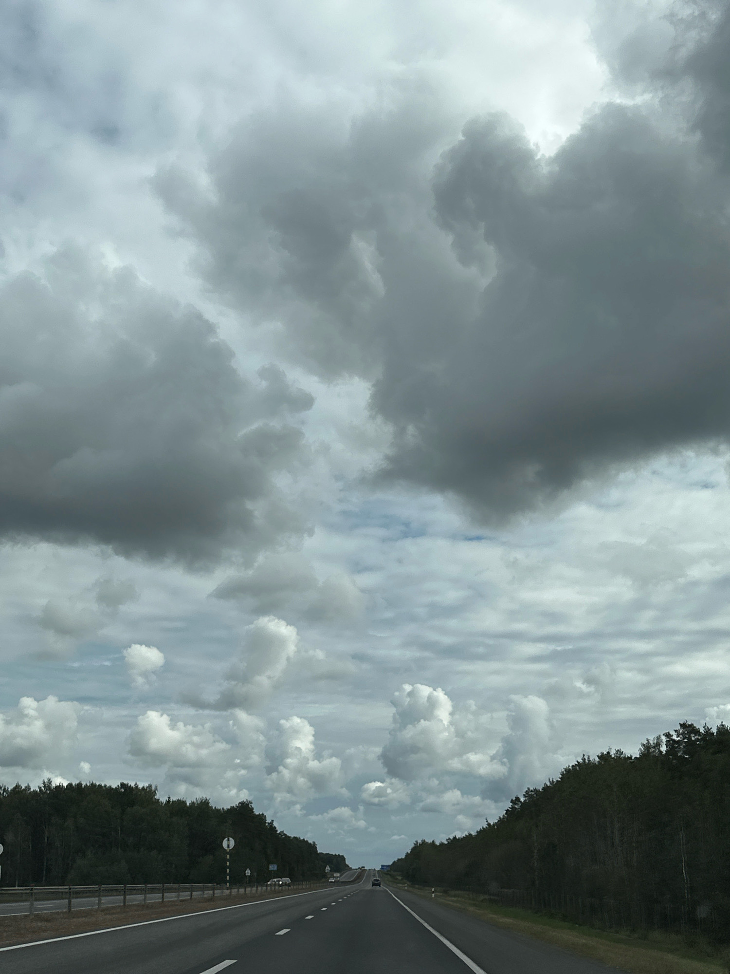 This is a forward-facing, eye-level, medium shot of a highway in Вяззеўскі сельскі Савет, Belarus, under a cloudy sky. The road is asphalt and has white lane markings. The highway is lined on both sides with trees. There are several cars on the road, traveling in the same direction as the camera. The sky is filled with large, dark gray clouds, with some patches of lighter clouds visible. The overall impression is one of a somewhat overcast or approaching stormy day.