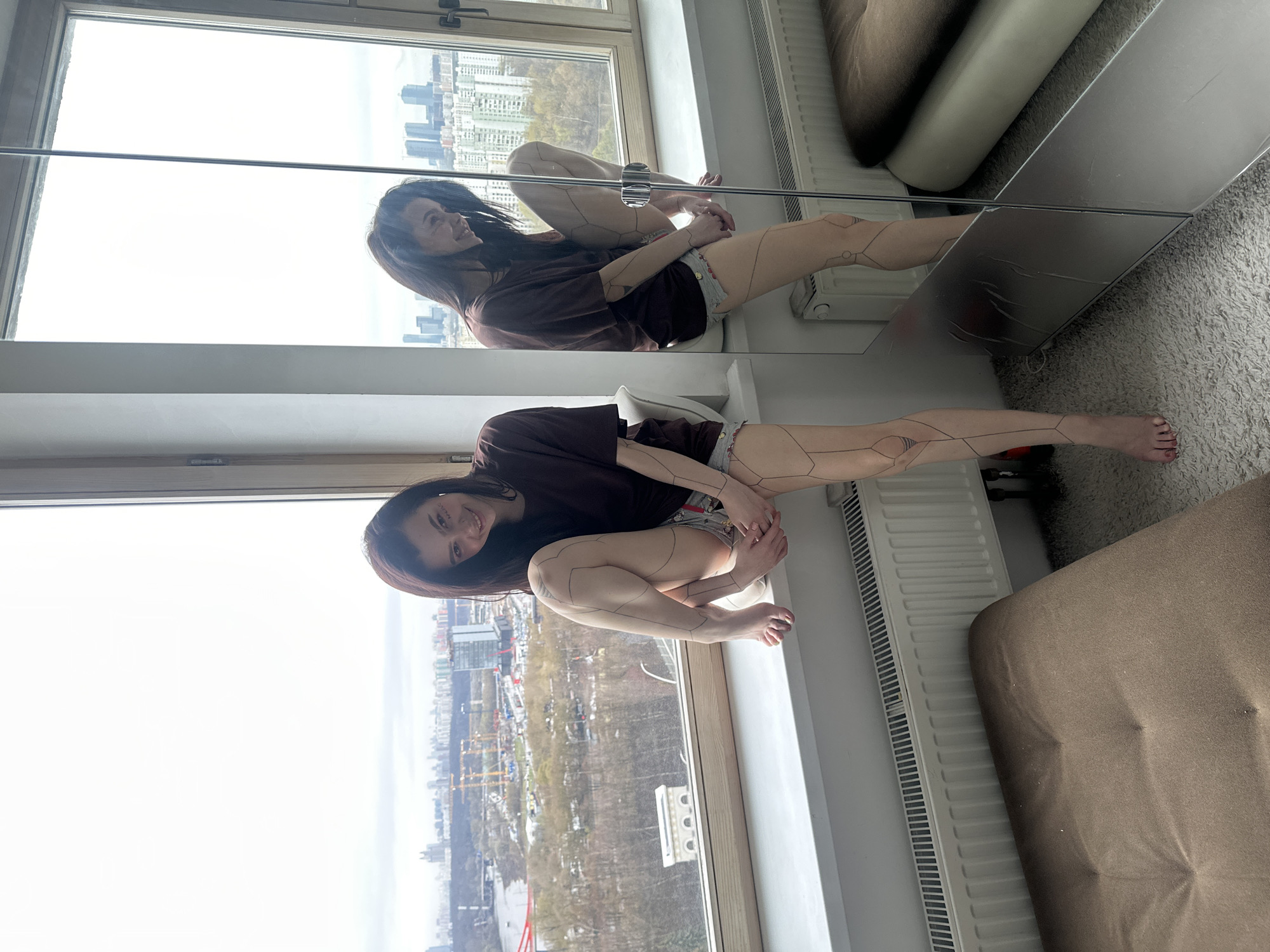 A young woman with dark hair is sitting on a window sill, looking at the camera and smiling. She is wearing a dark brown t-shirt and printed shorts. Her legs are decorated with geometrical patterns.
She is sitting in front of a large window, offering a broad view of Moscow, Russia. The cityscape is visible, including residential buildings, parks, and the Moskva River.
A mirror on the right reflects the scene, including the woman and the cityscape. The mirror also reflects a section of a radiator and a brown sofa. The room appears to be carpeted.