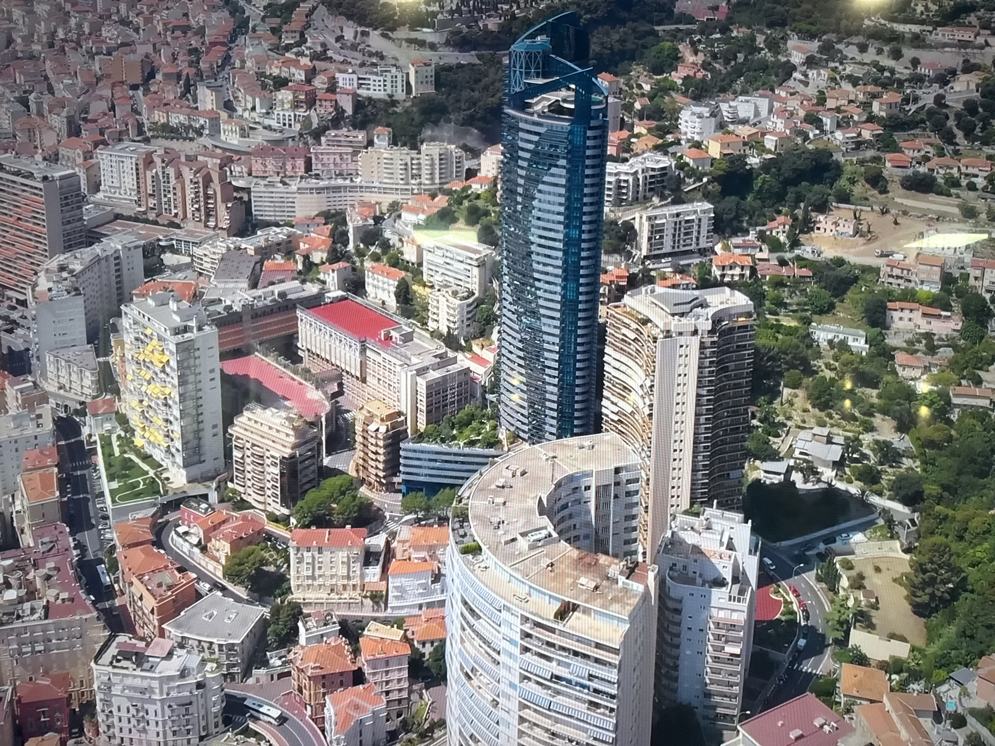 This is an aerial drone-style photograph of a dense urban environment, likely a city. The scene is dominated by numerous buildings of varying heights and architectural styles, interspersed with roads and patches of greenery. A prominent, tall, slender skyscraper with a distinctive blue-tinted glass facade stands out in the mid-ground, its upper sections featuring an unusual, multi-tiered design. Surrounding this skyscraper are many mid-rise apartment buildings, characterized by their red-tiled roofs and beige or light-colored facades. Some buildings are quite close together, forming narrow streets and alleyways, while others are more spread out, with gardens or small clearings visible. The terrain appears somewhat hilly, with roads winding through the landscape. The overall impression is one of a well-developed city with a mix of modern and older architectural elements. The image does not provide any visual cues to suggest the location is Dnipro, Ukraine; it is a generic urban landscape. There are no people visible in the photograph.