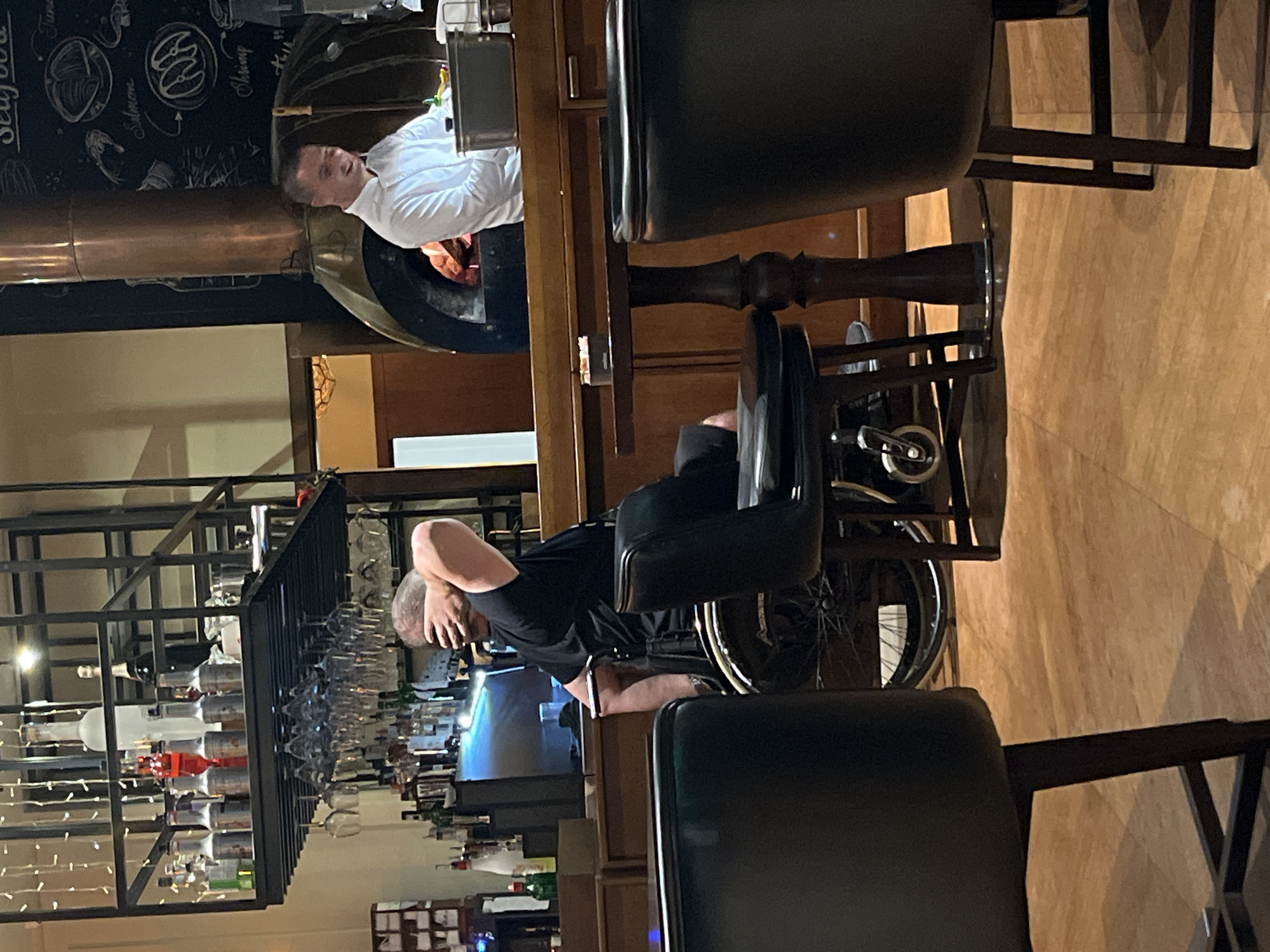 The image depicts an indoor scene at a restaurant in Budva, Montenegro. In the foreground, two black chairs with wooden legs are visible, one partially obscuring a wooden table with a dark, turned pedestal base. In the mid-ground, a person seated in a wheelchair is facing away from the camera, with their right arm raised to cover their face. They are wearing a black t-shirt. The wheelchair has a large spoked wheel. Behind the wheelchair, a bar area is visible with shelves stocked with bottles and glassware. A person in a white chef's jacket is standing behind a counter, smiling and looking to their left. A large, copper-hooded pizza oven is situated behind the chef. The environment suggests a lively, possibly upscale, dining establishment. The lighting is warm, with some artificial lights and what appears to be ambient light contributing to the atmosphere.