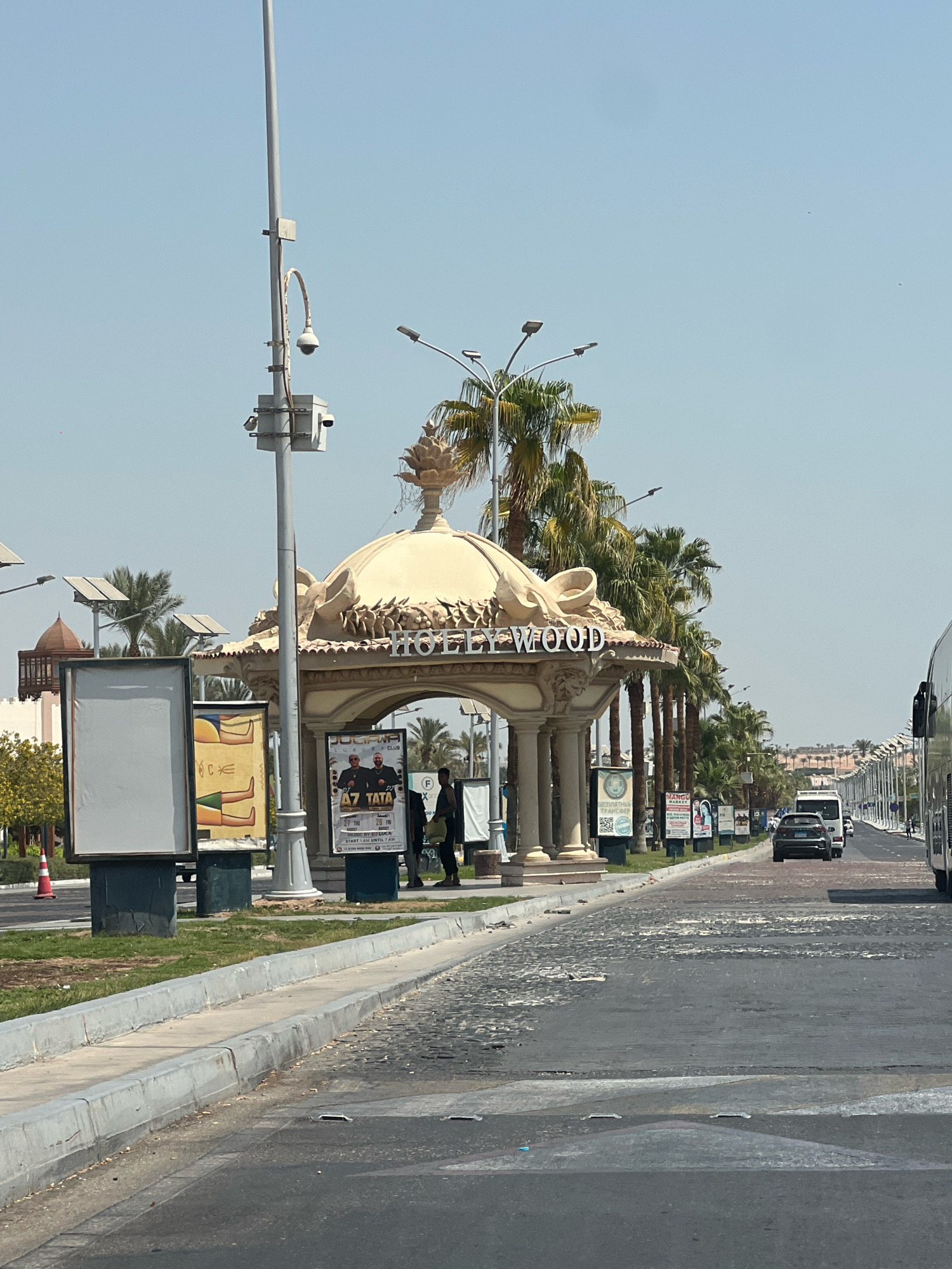 The image captures a street scene in Sharm El Sheikh, Egypt, with a prominent "HOLLYWOOD" sign visible on an ornate, beige-colored structure that appears to be an entrance or a pavilion. The structure has a domed roof with decorative elements and is supported by columns. Flanking the "HOLLYWOOD" sign are palm trees. In the foreground, to the left, are large, blank billboards and a lamppost with security cameras attached. A street with cars, including a dark-colored sedan and a white bus, proceeds into the distance. Several smaller billboards and posters are visible along the side of the road, some advertising events and businesses. Two individuals are standing near the "HOLLYWOOD" structure, one near a poster and another further back. The sky is clear and blue, indicating a sunny day. The road surface in the foreground appears to be cobblestone or a similar textured material that is partially worn or repaired. In the background, a row of streetlights and buildings can be seen stretching into the distance, suggesting a developed area within the city.