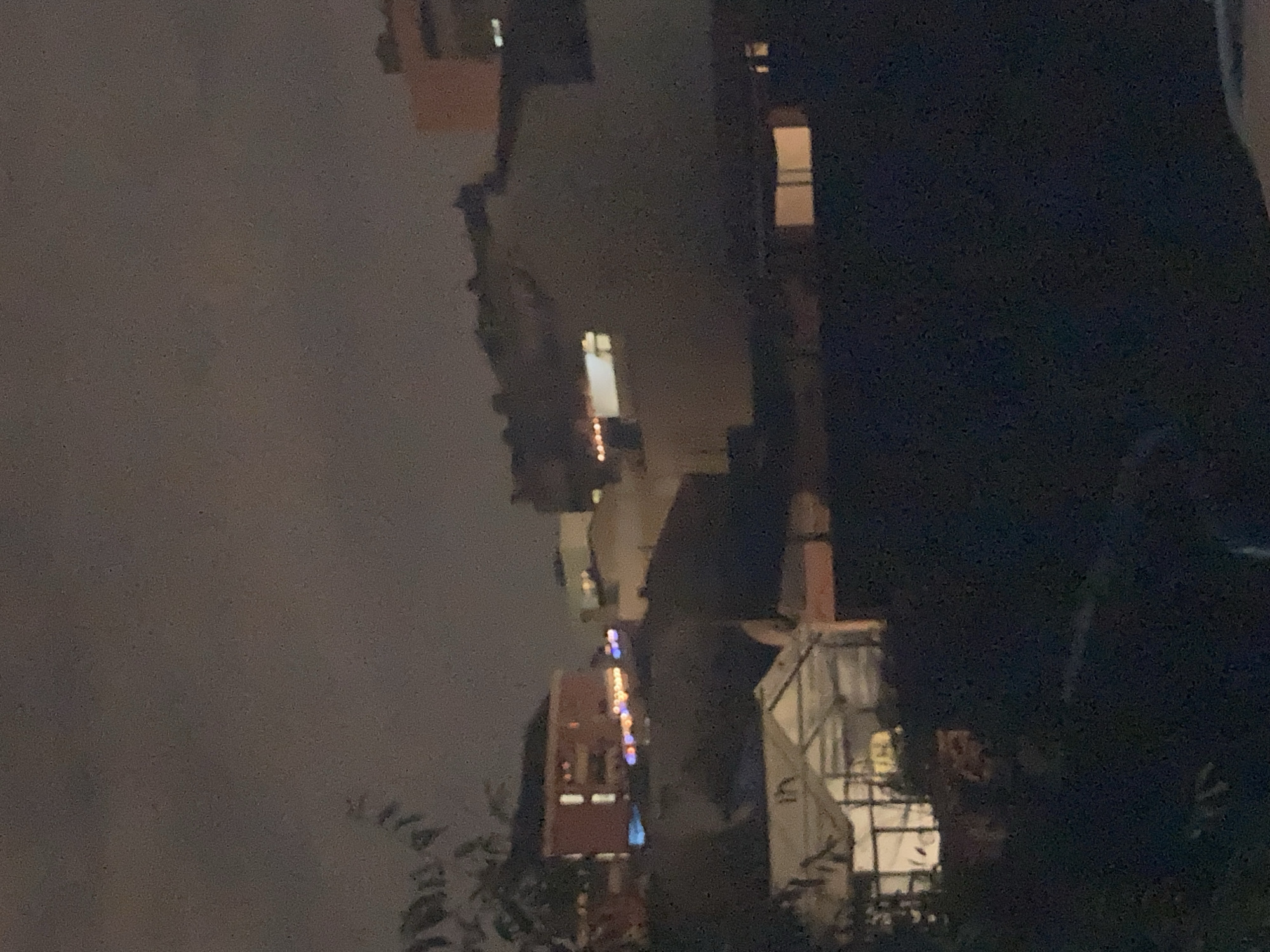 This is a nighttime exterior shot of a residential area in Sabadell, Spain. The image shows several buildings with illuminated windows and some decorative lights. In the foreground, there is a dark, silhouetted area with some foliage, possibly a tree branch, on the left side. In the midground, a greenhouse-like structure with internal lighting is visible. Behind it, a multi-story building with red brick and visible windows is illuminated with orange and blue lights along its length. Further back, other buildings with lit windows are discernible against the dark sky. The overall lighting suggests it is after sunset or before sunrise.