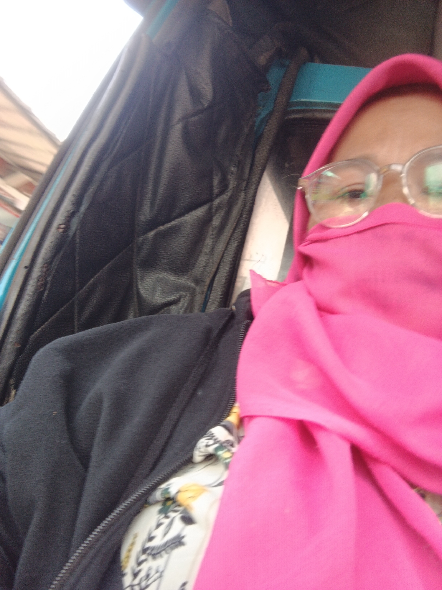 A close-up, slightly blurred, over-the-shoulder selfie shows a person wearing a bright pink hijab and glasses, with their face partially covered by the hijab. The person is wearing a black jacket over a white shirt with a floral pattern. The background includes a black padded interior, possibly of a vehicle, with a teal blue element visible. The scene is set in Kebonmanggu, Indonesia.