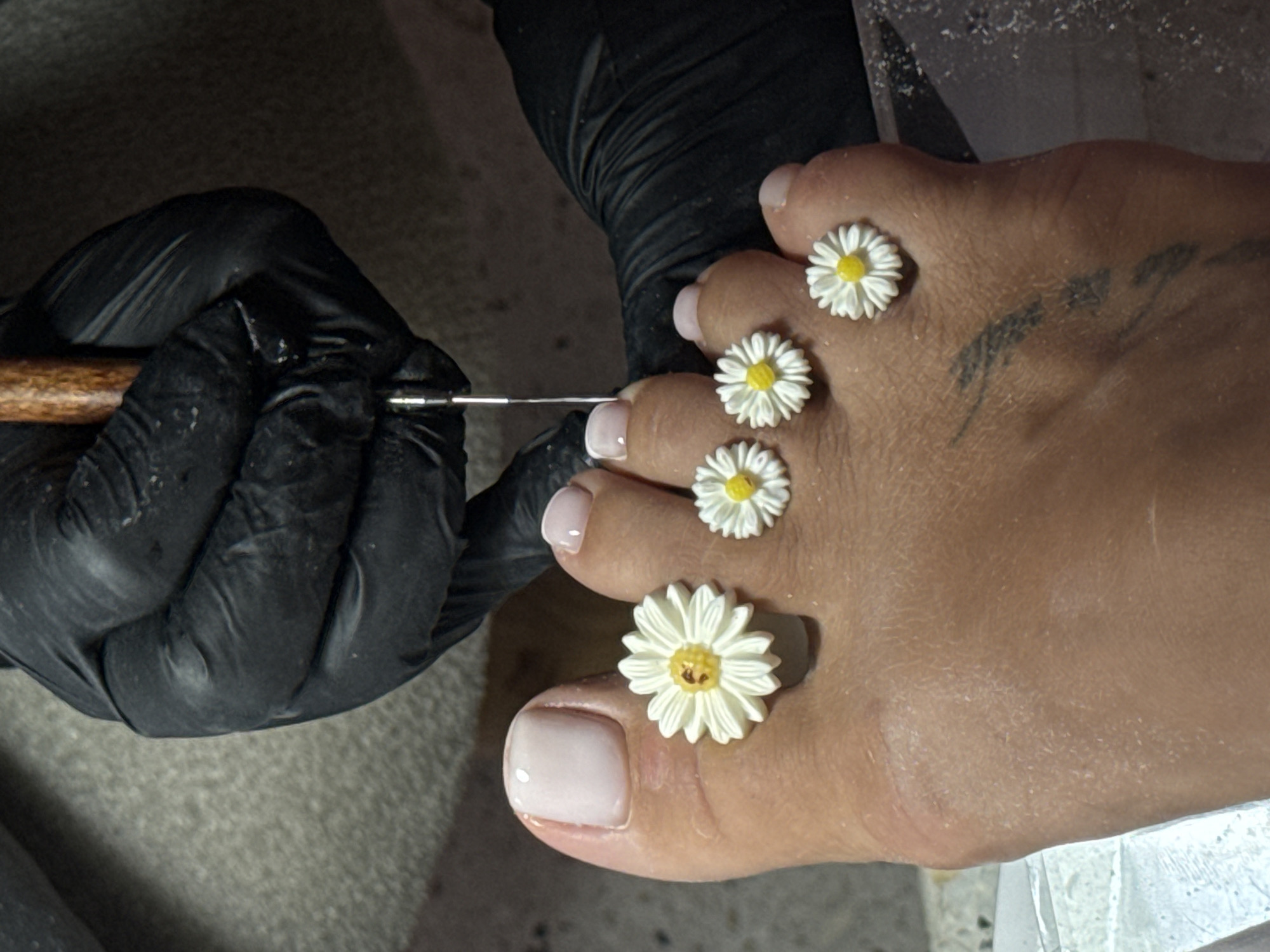 This close-up image captures a detailed foot care or nail art session taking place in North Kuta, Indonesia. The main subjects are a client's foot and a service provider's gloved hand.
The foot, positioned horizontally in the frame, features four delicate, three-dimensional daisy decorations with white petals and yellow centers, arranged along the top surface of the foot, between the toes and the ankle. The toenails are neatly trimmed and painted with a glossy, light pink or nude polish. A faint, dark tattoo, appearing to be script or abstract lines, is visible on the top of the foot, towards the ankle. Some fine, light-colored dust or debris is scattered across the foot and surrounding surfaces, possibly from nail preparation.
A hand, encased in a black nitrile or latex glove, is holding a slender nail art tool with a wooden handle and a metallic tip. The gloved hand is positioned on the left side of the frame, meticulously working near the big toe, suggesting the application of fine details or embellishments.
The setting appears to be an indoor environment, likely a nail salon or spa, with a dark, textured material (possibly a towel or mat) serving as a background for the gloved hand and a lighter, reflective surface at the bottom right. The lighting is focused, highlighting the activity. No specific time of day or weather cues are discernable, and there is no visible text in the image.