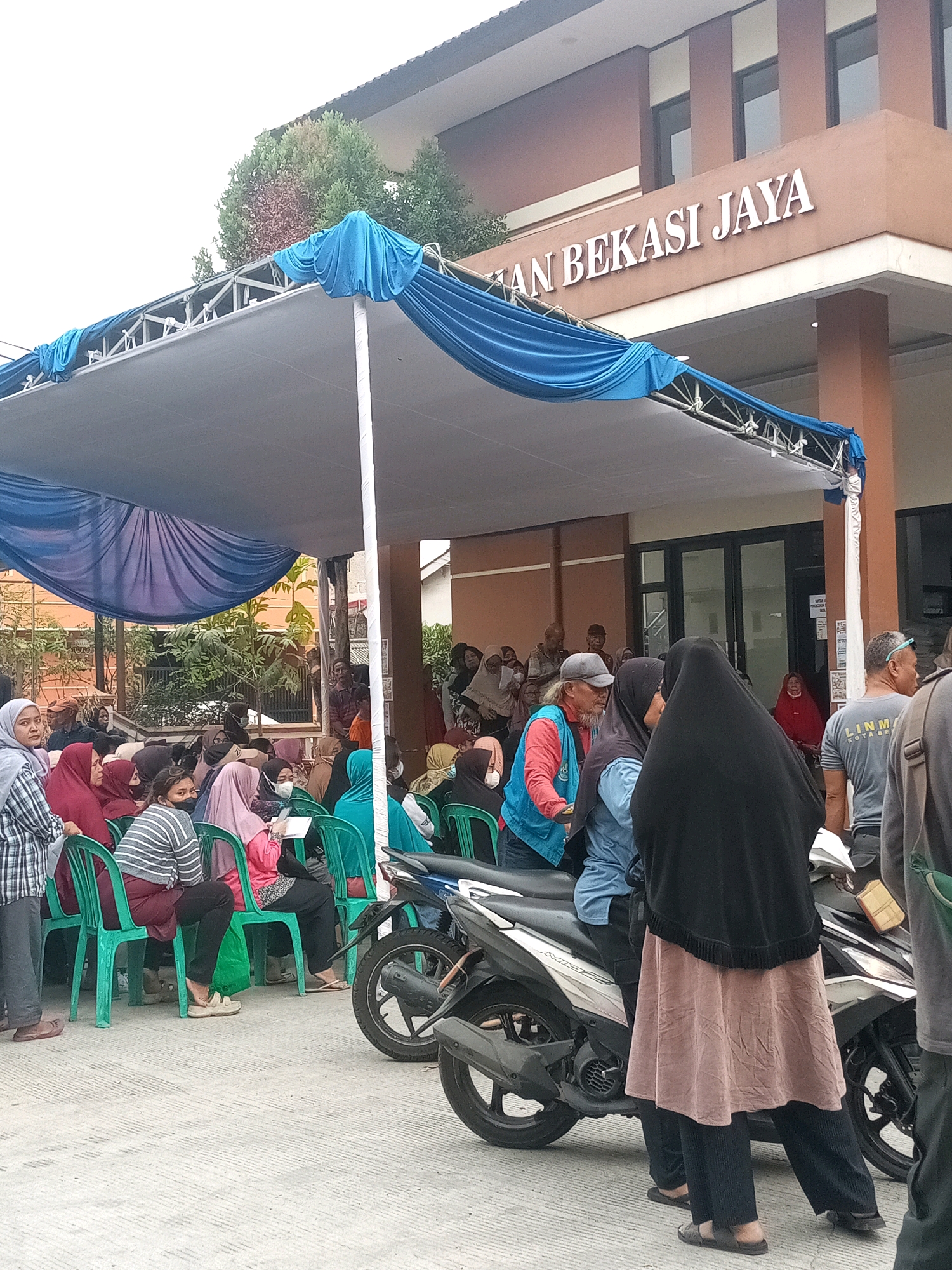The image depicts an outdoor scene in front of a building with prominent signage reading "AN BEKASI JAYA". A large, blue-draped canopy shelters a gathering of people seated on green chairs. Many individuals are wearing headscarves, suggesting a cultural context common in Indonesia. A motorcycle is parked in the foreground. The overall atmosphere suggests a public event or service being held outdoors, likely in the city of Bekasi, Indonesia, given the building's signage. The people appear to be waiting or observing, with some interacting with each other. The environment is paved, and the building behind the canopy has a brown facade.