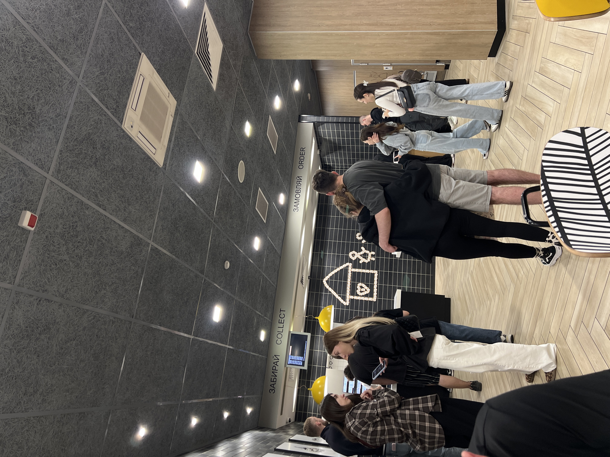 This media file depicts a group of people standing in a brightly lit indoor setting, likely a fast-food restaurant. The scene is captured from a high angle, looking down at the individuals and the environment.
The primary subjects are several young adults, both male and female, who appear to be in a queue. They are dressed in casual, modern attire, including jeans, t-shirts, hoodies, and sneakers. Some individuals are interacting with their phones, while others are looking towards a counter area. One couple is embracing.
The setting includes a modern interior with a dark tiled wall featuring a grid pattern and a white sign with text in Cyrillic. Above the counter, there's a black and white tiled wall with some decorative elements, including a white house outline made of what appears to be food items. Two bright yellow balloons are also visible. The ceiling is a textured acoustic panel with recessed lighting and air conditioning vents. A wooden floor with a parquet pattern is visible. A chair with a black and white striped seat is also in view.
The visible text includes "ЗАМОВЛЯЙ ORDER" and "ЗАБИРАЙ COLLECT" on white signs, indicating an ordering and collection point.
The overall impression is that of a casual social gathering or a group of friends in a public eating establishment. The lighting suggests it could be daytime, but without external views, it's difficult to determine the time of day or weather. The emotions conveyed are neutral, with a sense of everyday activity. The location context is identified as Ternopil, Ukraine.