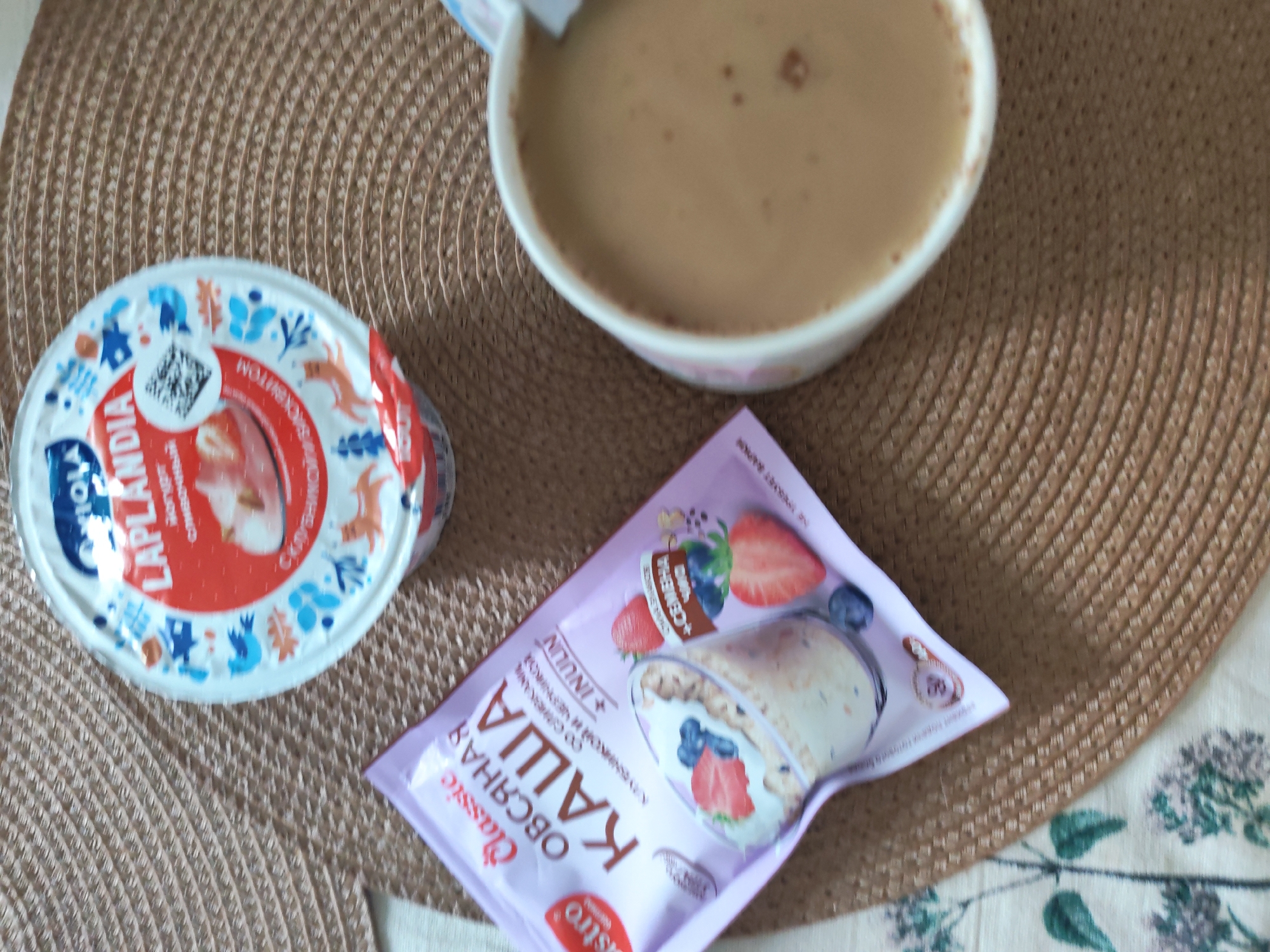 The image shows a table setting with breakfast items. There is a cup of coffee, a container of Laplandia yogurt, and a packet of Bistro Classic oatmeal. The items are placed on a woven placemat, with a portion of a floral-patterned tablecloth visible in the lower right corner. There is no indication in the image that suggests this is in Izhevsk, Russia.