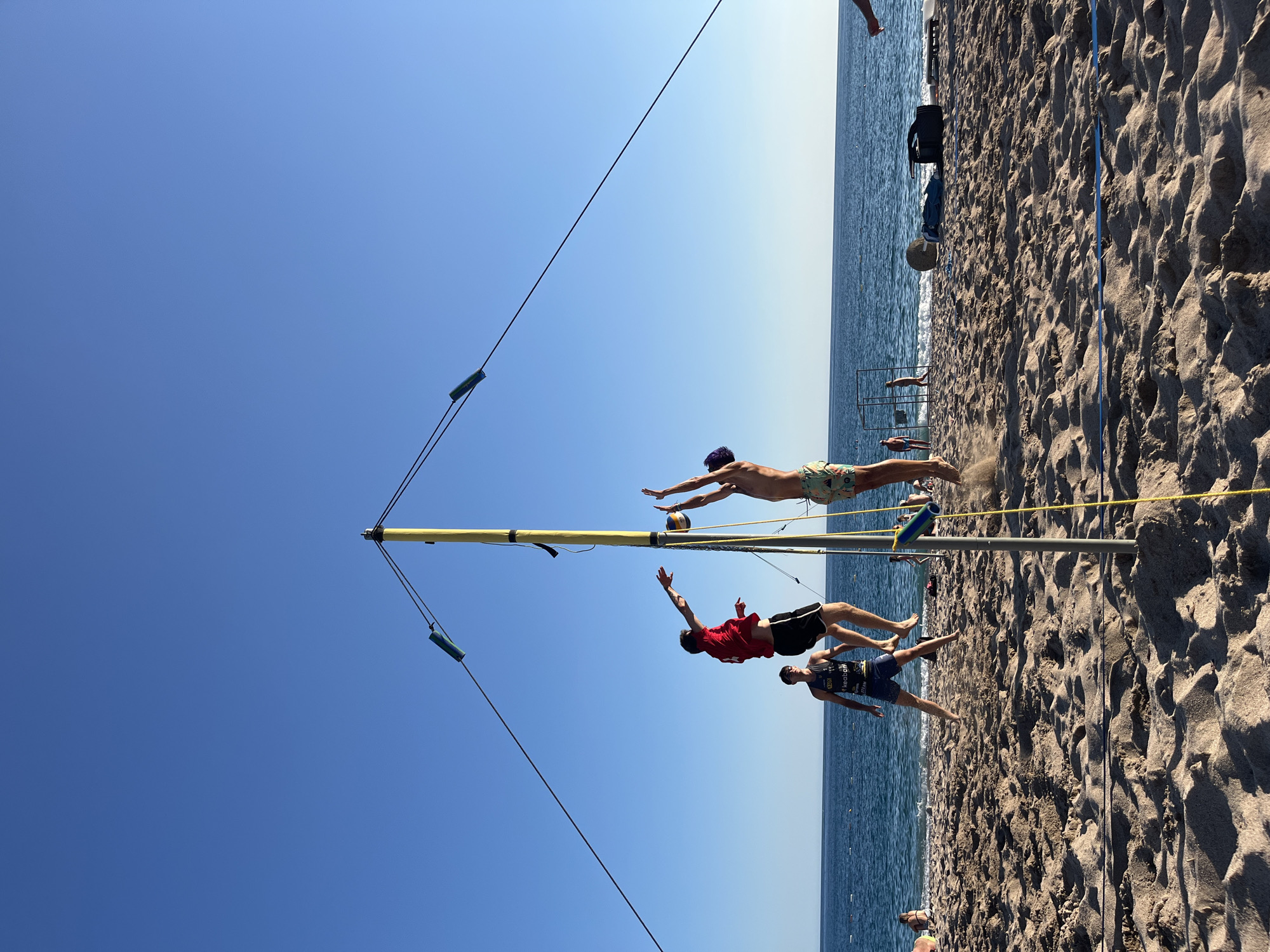 The image shows a beach volleyball game on a sunny day at Zankovići, Montenegro. The sea is visible in the background, with a clear blue sky above. The sand is a light brown, with footprints and waves imprinted on it. A volleyball net is set up on the beach, with three men actively playing.
The subjects are three men engaged in a volleyball match. The man in the middle is jumping to hit the ball, with his body extended in mid-air. He is wearing a red t-shirt and black shorts. To his left, another man is also jumping, reaching for the ball with his right hand. He is wearing patterned swim trunks. To the right of the man in the red shirt, a third man is positioned lower, appearing to be ready to receive the ball or make a defensive move. He is wearing a dark tank top with text and dark blue shorts.
The activity taking place is a dynamic moment in a beach volleyball game, likely a spike or a block attempt. The players appear focused and athletic. The time of day is clearly daytime, with bright sunlight casting shadows on the sand and highlighting the players' movements. The weather is fair and sunny, as indicated by the clear sky and the activity on the beach.
Notable details include the sand kicking up around the jumping player, suggesting a powerful move. The volleyball net structure is visible, with yellow and blue elements. A yellow pole supporting parts of the net structure is prominent. There are also some people in the background on the beach, enjoying the sunny weather. The overall mood is energetic and sportive. Some text is visible on the tank top of the man on the right, but it is not clearly legible.