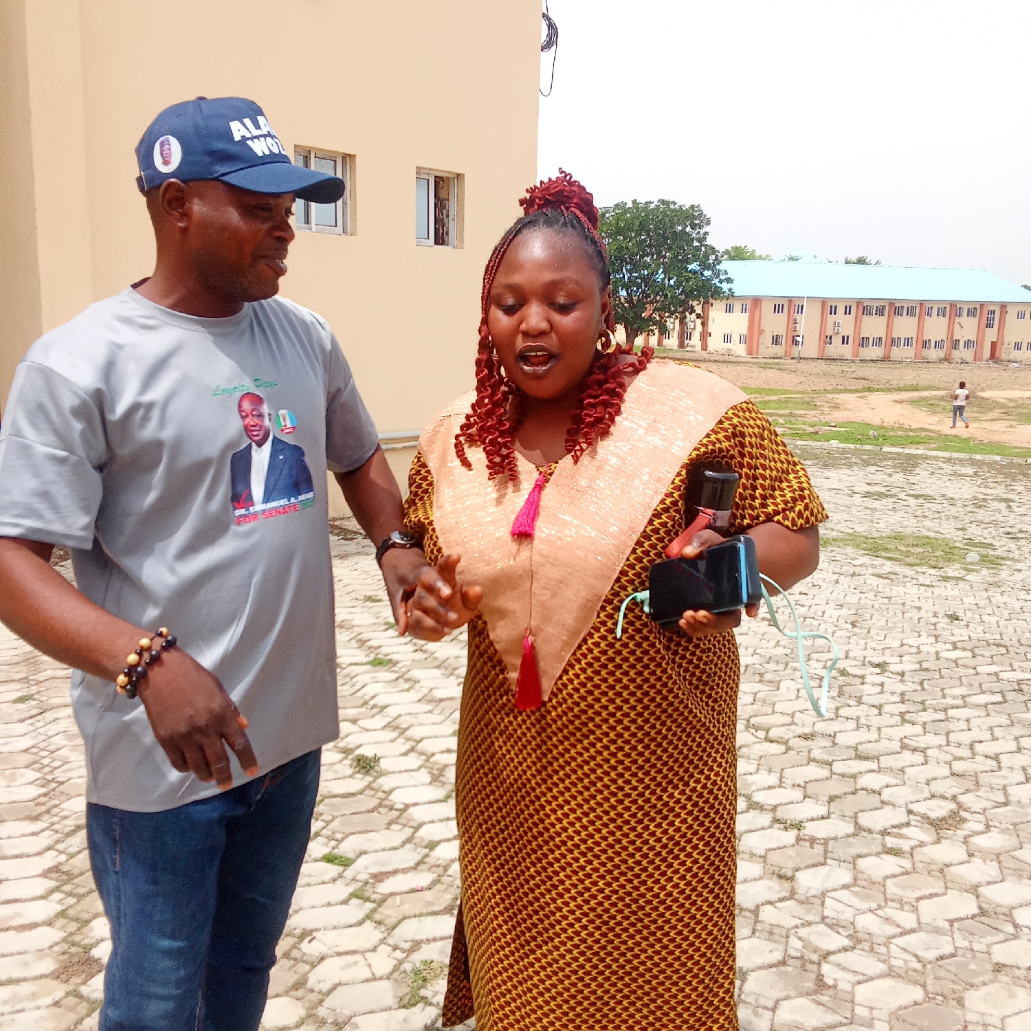 This daytime image, set in Akwanga, Nigeria, captures a candid interaction between a man and a woman outdoors under clear, sunny skies.
On the left, a man is seen wearing a grey t-shirt, blue jeans, and a blue baseball cap. His t-shirt prominently displays a portrait of a man with the text "DR. ADEWUNMI ALAO FOR SENATE 2023" and the phrase "Logun Eje." The cap features the partial text "ALA WO" and a small circular image of a face. He also wears a beaded bracelet on his right wrist. He is looking towards the woman, appearing engaged in conversation with a pleasant, slightly smiling expression.
On the right, a woman is dressed in a vibrant patterned gown in shades of brown and gold, featuring a lighter beige overlay on the bodice with a decorative red tassel. Her hair is styled in distinctive red/auburn braids, and she wears large hoop earrings. She is holding a black smartphone and what appears to be a power bank or portable speaker, connected by a light blue cable. Her mouth is open, suggesting she is speaking or singing animatedly.
The background features light-colored buildings, possibly an institutional or campus setting. A building with a light blue roof is visible in the distance, and a large tree stands between the structures. The foreground is a paved area with hexagonal-patterned blocks, transitioning to dry, unpaved ground further back where a lone figure walks in the distance. The political branding on the man's attire suggests a context of public engagement or a campaign event.