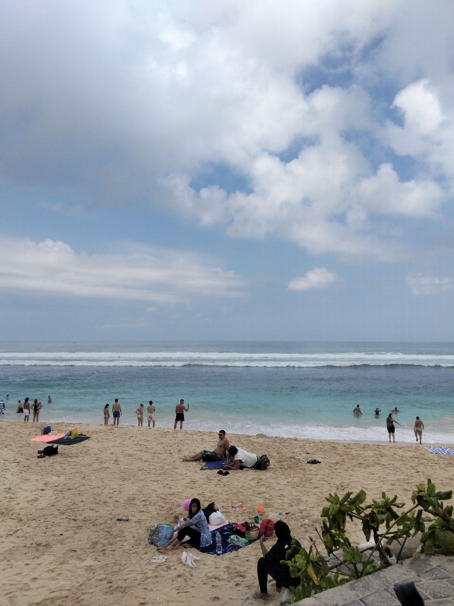 The image depicts a sandy beach with several people in and near the ocean. In the foreground, a woman in black clothing is sitting on the sand, with a black bag beside her and some green foliage in the lower right corner. Further up the beach, a group of people are relaxing on towels, including a man in green shorts and a woman lying down. A few beach towels and bags are scattered on the sand. In the background, the ocean has moderate waves, and several people are visible wading or swimming in the water. The sky is partly cloudy. The scene appears to be located on a beach in Kuta Selatan, Indonesia, a popular tourist area known for its beaches.