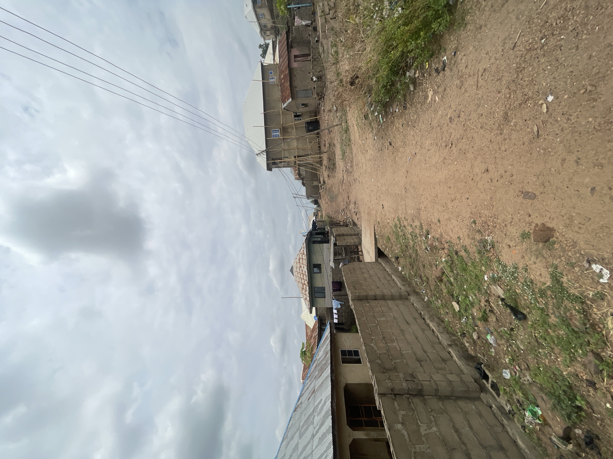 The image depicts a dirt road lined with buildings in Lokoja, Nigeria, under a cloudy sky. The road is uneven and strewn with litter, with vegetation growing along the edges. The buildings are a mix of completed and under-construction structures, primarily made of concrete blocks. Some have corrugated metal roofs. Electrical wires are visible overhead. No people are visible in the scene.