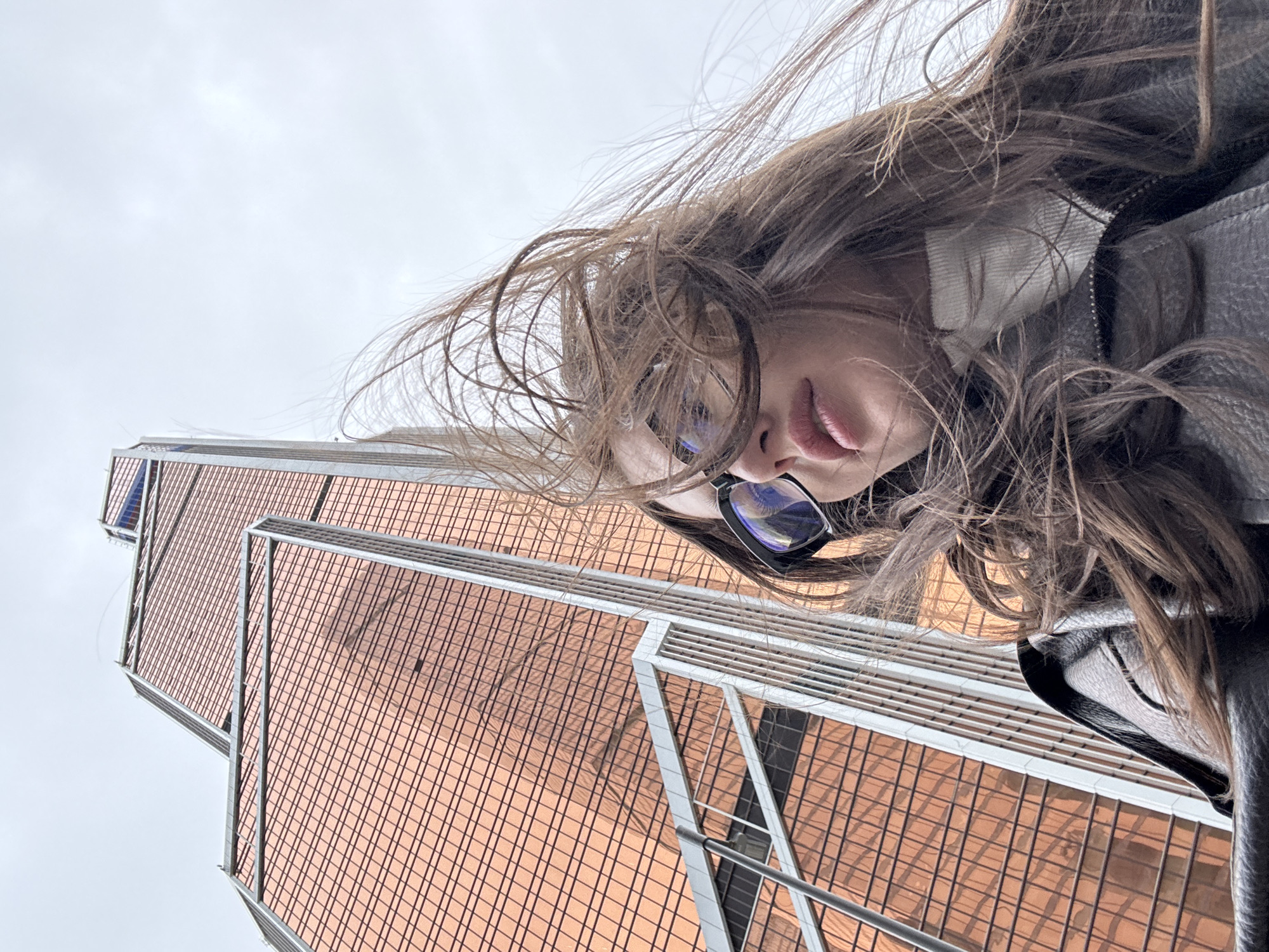 A woman wearing glasses and a leather jacket is seen in a low-angle shot with her hair blowing in the wind. Behind her is the Mercury City Tower, a skyscraper in the Moscow International Business Center (Moscow-City), Russia. The building's facade is made of orange-tinted glass and is framed by metal supports. The sky is overcast.