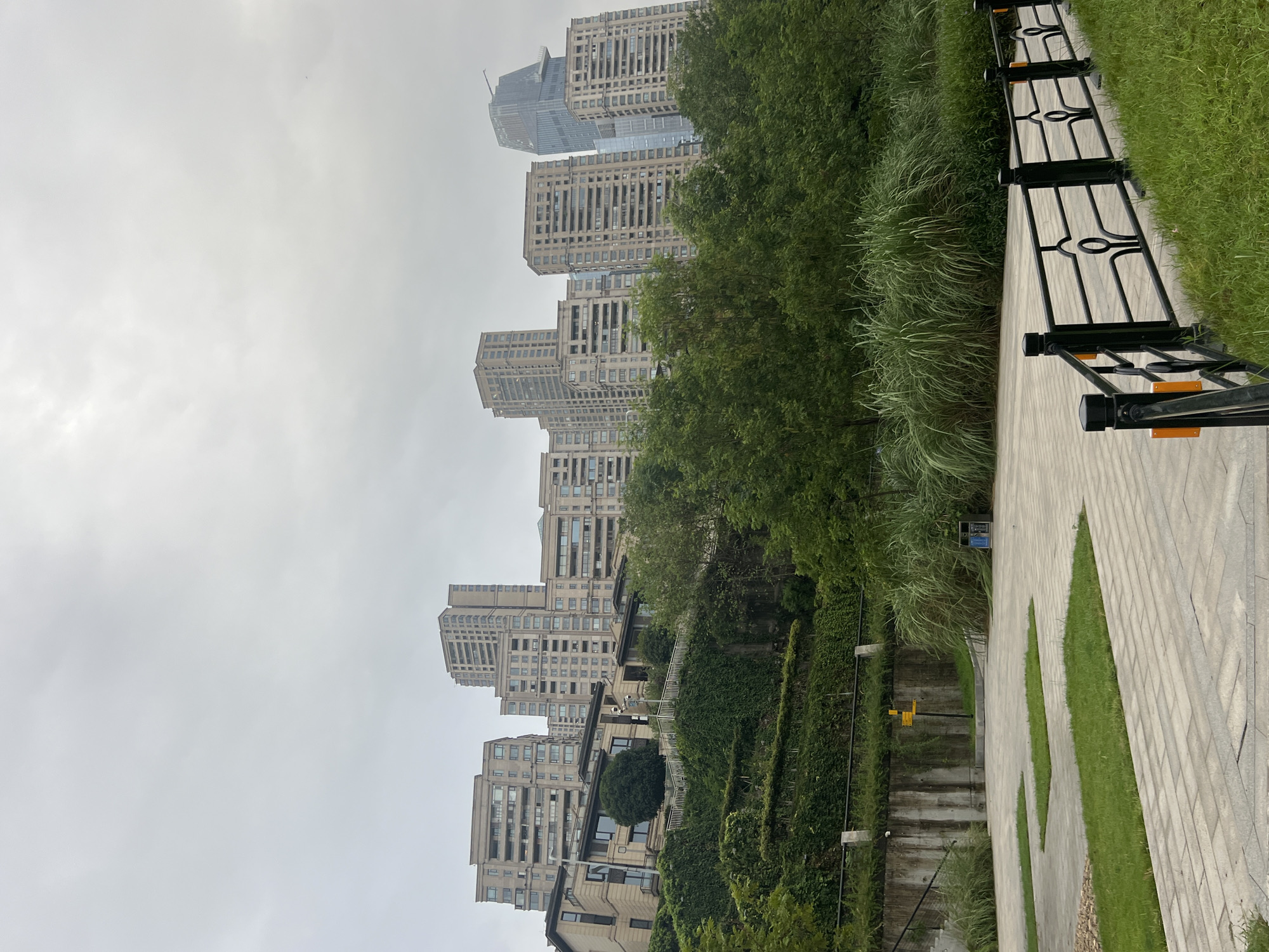 The image depicts a residential area with several high-rise apartment buildings and some lower-rise structures built on a hillside. The buildings are modern and appear to be made of concrete and glass. Lush green vegetation, including trees and vines, covers the slopes of the hill. In the foreground, there is a paved pathway with decorative grass patches and a black metal fence with ornate railings. A trash can is visible near the pathway. The sky is overcast, suggesting a cloudy day. The scene appears to be in a city, and the mention of "江北城街道, China" places it in that specific location. There are no people visible in the image. The overall impression is one of a well-maintained urban residential landscape.