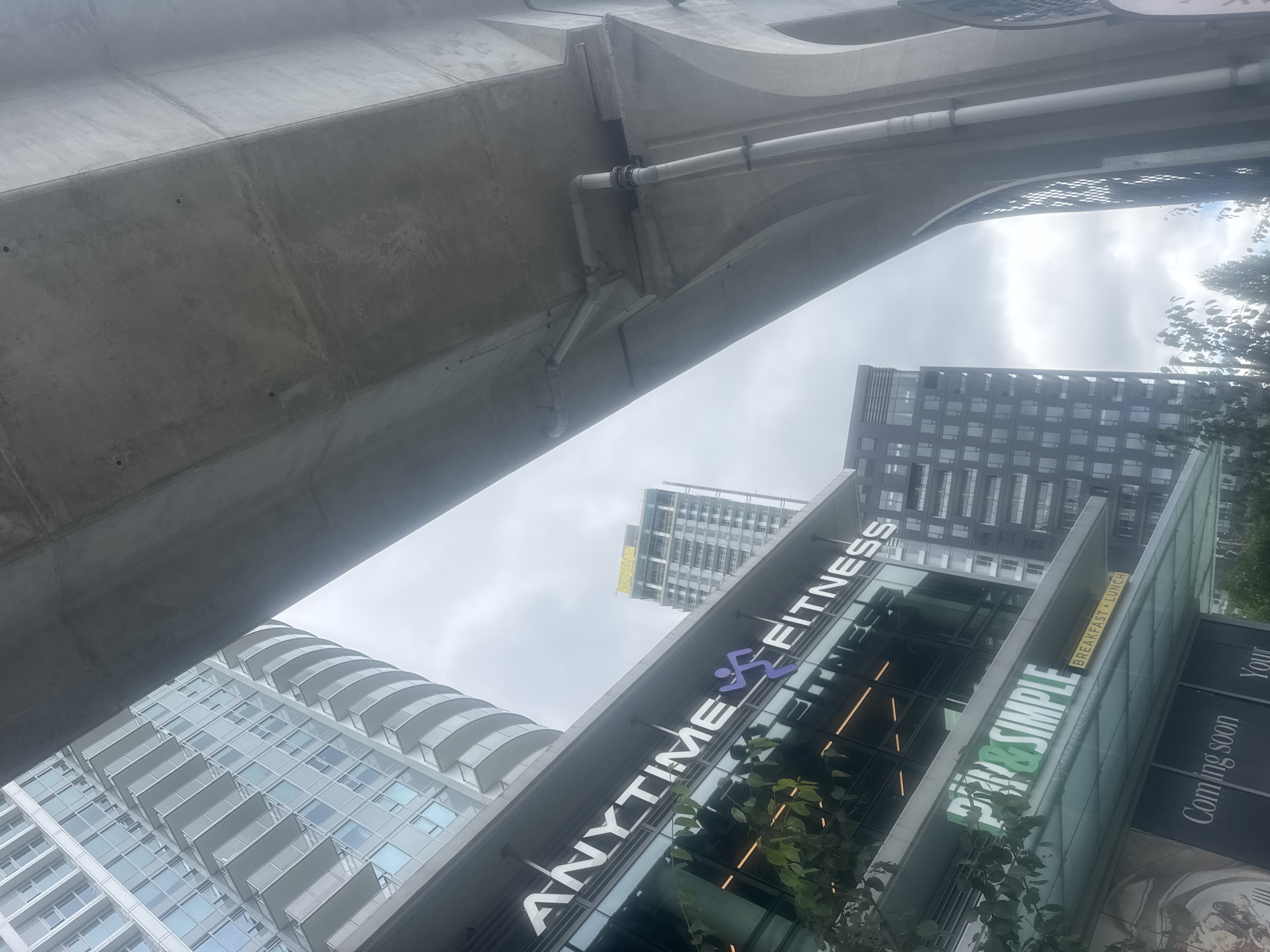 The image shows an outdoor view of the Lougheed Highway overpass in Coquitlam, Canada. In the foreground, there is a building with the signs "ANYTIME FITNESS" and "PIN&SIMPLE BREAKFAST&LUNCH". A modern, tall, glass-fronted residential building with balconies is visible to the left, and a darker, more densely windowed residential building is seen to the right and behind the "ANYTIME FITNESS" sign. The concrete structure of the overpass dominates the upper portion of the image, with visible pipes and conduits attached to its underside. The sky is overcast, suggesting a cloudy day.