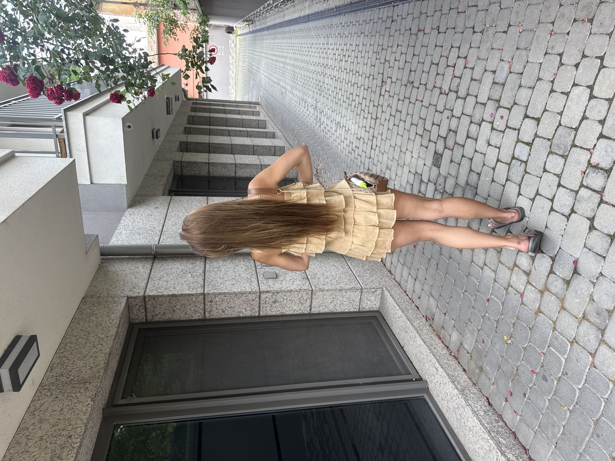 A daytime image, rotated ninety degrees, captures a person from behind, standing on a paved outdoor walkway in an urban setting. The individual has long, straight, light brown hair flowing down their back and is wearing a light beige or tan dress with ruffled layers. They carry a small, patterned shoulder bag, with a yellow pen or marker and a white tube partially visible inside. Their legs are bare, and they are wearing flat, dark sandals.
The walkway is composed of grey interlocking cobblestones, with scattered pink petals adding subtle splashes of color. To the left (when viewed upright), a modern building features light-colored walls and grey granite-like accents. A set of outdoor steps ascends beside the building, and a large, dark rectangular window or glass panel is visible at ground level. A minimalist rectangular light fixture is mounted on the wall.
Above the building, green foliage with vibrant dark red roses and leaves is present. The paved path extends into the distance, suggesting a pedestrian area. The scene is illuminated by natural light, indicating daytime with soft, possibly overcast, conditions as there are no strong shadows. The location is identified as Plovdiv, Bulgaria. No legible text is visible in the image.
