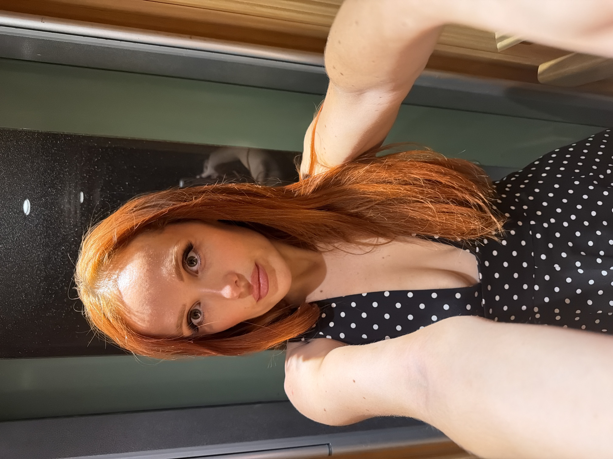 A woman with long red hair, light skin, and wearing a black polka-dot dress takes a selfie. Her left hand holds her hair near her neck, while her right hand holds the camera. The background includes a counter, cabinet doors, and a light-colored ceiling. The location is not discernible from the image.