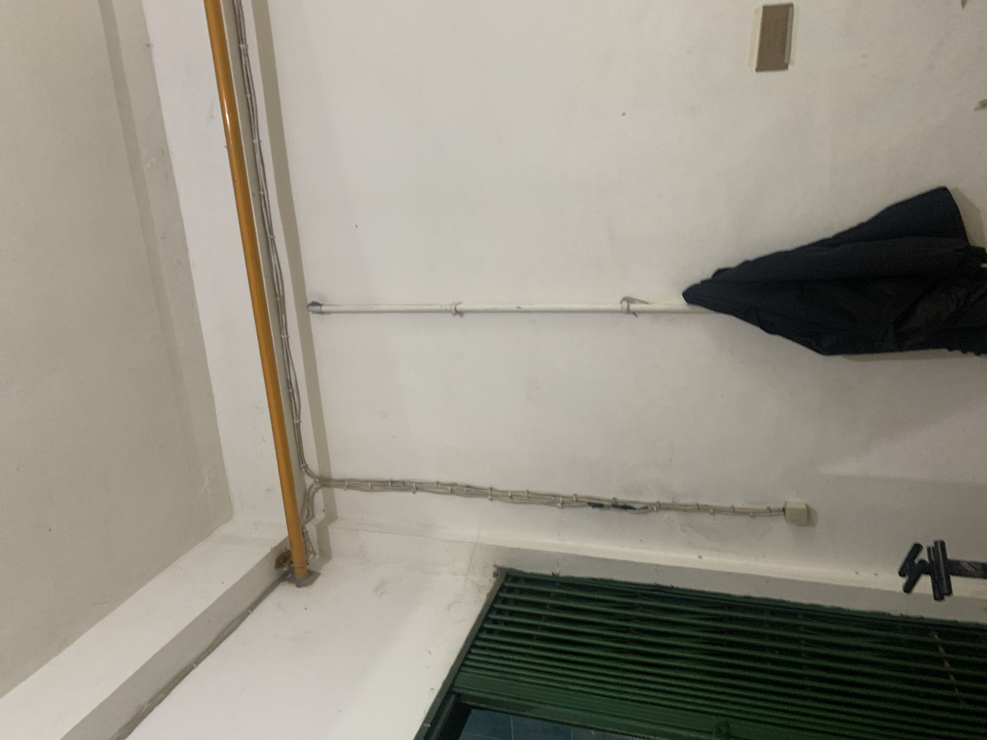 This image depicts an interior scene, likely a utility area or storage space, in Palembang, Indonesia. A yellow conduit runs horizontally along the ceiling, with several electrical wires and pipes running parallel to it. To the left, a vertical arrangement of electrical wires is attached to the wall, leading down to an electrical outlet. A green, slatted metal door or gate is partially visible on the far left. On the right side of the image, a white pipe runs vertically down the wall, with a dark garment, possibly a jacket, hanging from it. A small, rectangular object, possibly a light switch or signage, is mounted on the wall to the right of the hanging garment. The walls and ceiling appear to be painted white, showing some signs of wear and tear. The overall impression is one of a functional, possibly industrial or service-oriented, space.