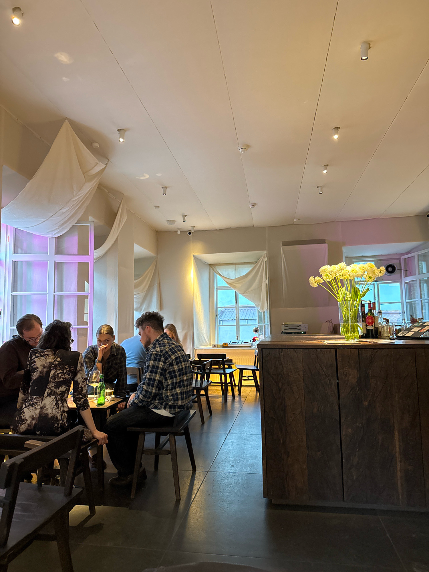 The image shows the interior of a restaurant in Moscow, Russia. A group of people are seated at a table in the foreground. A man in a plaid shirt is on the right, facing away from the camera. To his left, a woman with dark hair and a patterned dress is also seated. In front of them, a man with glasses and a dark shirt is seated, and next to him, a woman with light brown hair. They appear to be engaged in conversation or dining. The restaurant has a modern, minimalist aesthetic with white draped fabric hanging from the ceiling. There are large windows with white frames, and some have a purple hue. A bar area is visible on the right side of the image, with bottles and a vase of yellow flowers. The flooring is dark.