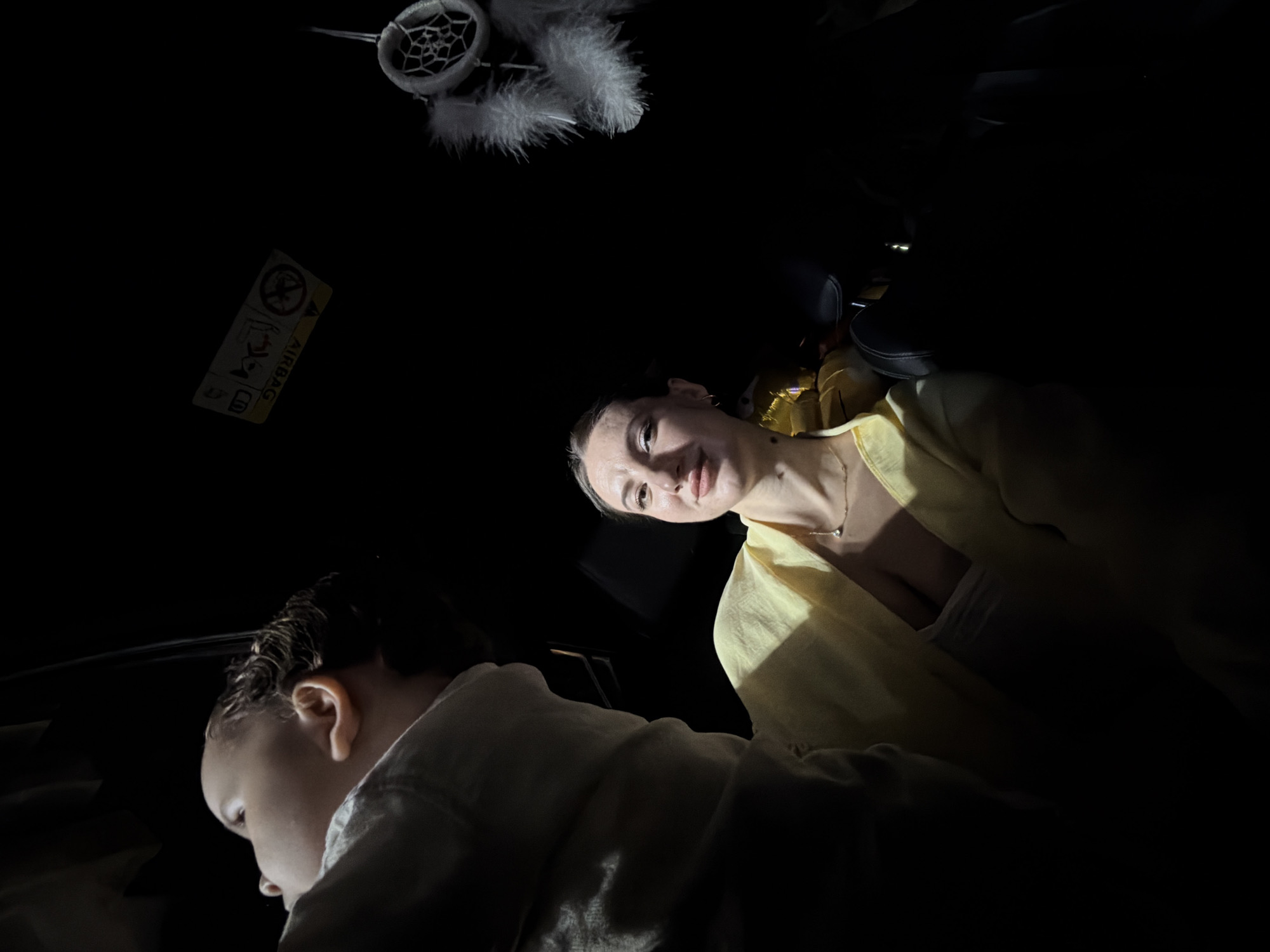 In the interior of a vehicle, a woman is positioned in the foreground, facing the camera with a slight smile. She is wearing a yellow top and a necklace. To her left and slightly behind her, a child's head is visible, looking upwards and away from the camera. The vehicle's interior is dark, with an "AIRBAG" warning sticker visible on the ceiling. A small dreamcatcher with white feathers hangs from above. The scene appears to be captured at night, possibly on a street in 6th of October City, Egypt.