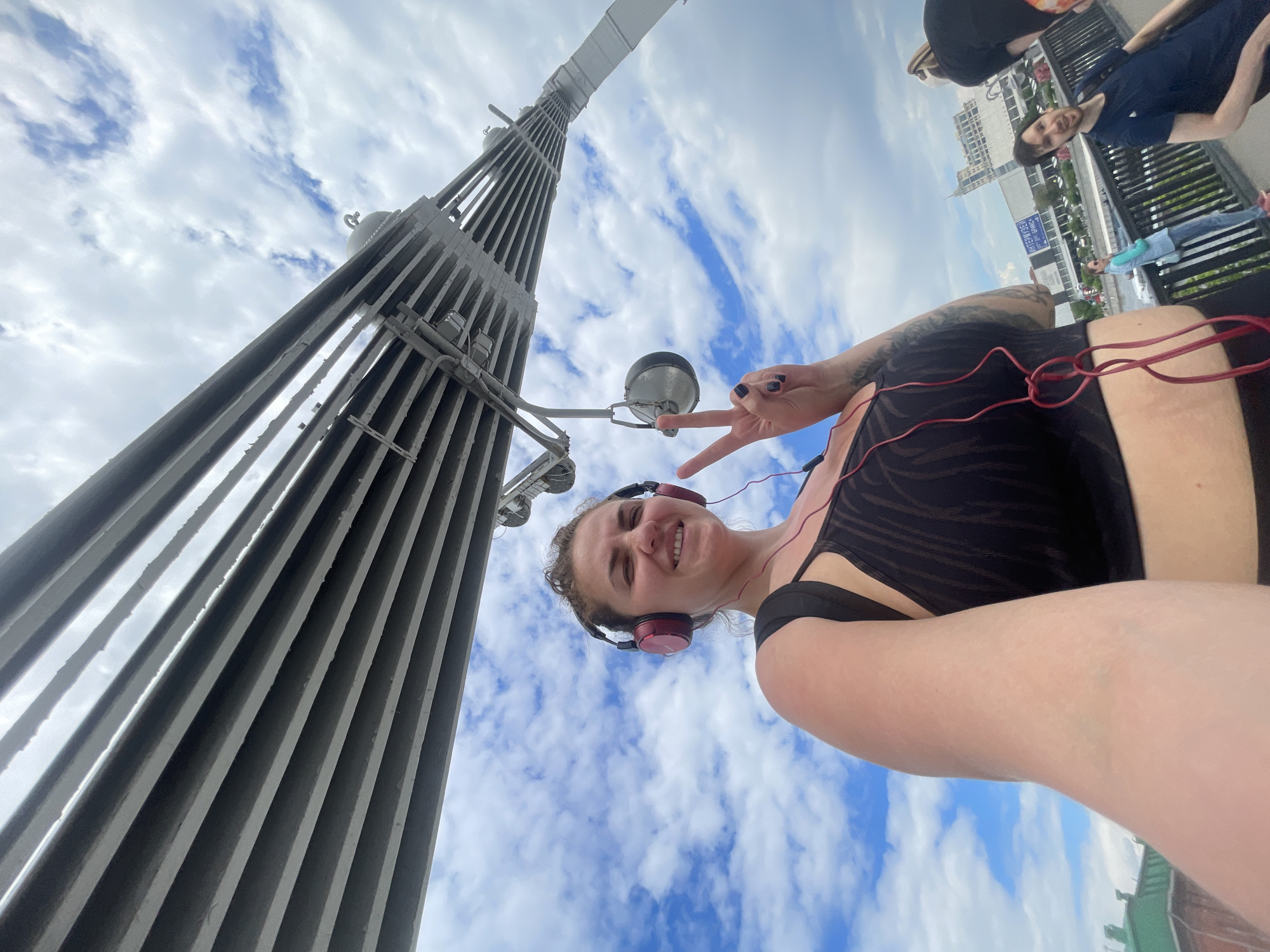 The image is taken from a low angle, looking up at a metal structure that appears to be a tower or monument. A woman is posing in the foreground, wearing headphones and making a peace sign with her hand. Other people are visible on a bridge or walkway. A building with signage in Cyrillic script is in the background. The sky is partly cloudy. This photo was taken in the city of Moscow, Russia.