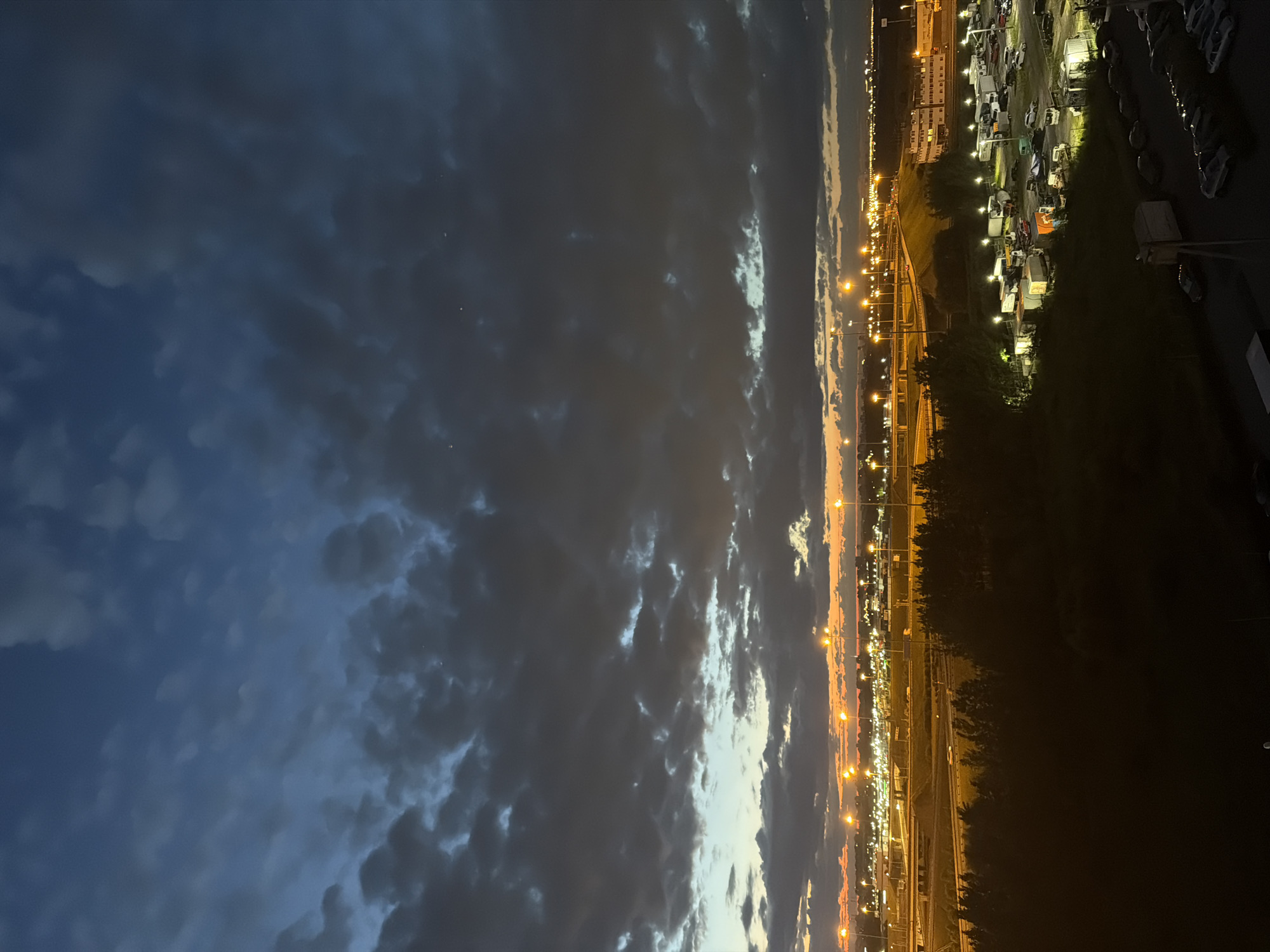 An aerial, wide shot captures a twilight scene with a dark, cloudy sky. The horizon is illuminated by a line of orange streetlights and distant city lights. In the foreground, dark trees obscure a large, partially lit parking area filled with numerous trucks, trailers, and cars. The scene appears to be an industrial or logistical area on the outskirts of a city, with a highway visible in the mid-ground.