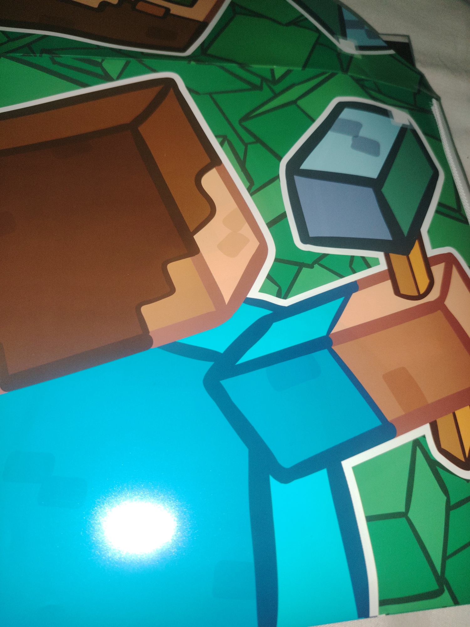 The image shows a close-up of a Minecraft-themed graphic. The graphic features blocky, pixelated characters and objects typical of the game, rendered in bold outlines and flat colors. Dominant colors include brown for blocky textures, bright blue for character clothing or body parts, and various shades of green for the background, which appears to be a stylized representation of grass or foliage with geometric patterns. The lighting in the image suggests it might be indoors, with a bright, possibly artificial, light source causing a glare on the blue surface. The image is a partial view, showing portions of two figures, one with a brown blocky arm and the other holding a blue and grey cube object. The overall impression is of a colorful and stylized depiction of elements from the video game Minecraft. The image is not situated in Xonobod City, Uzbekistan; it is a generic graphic representation.