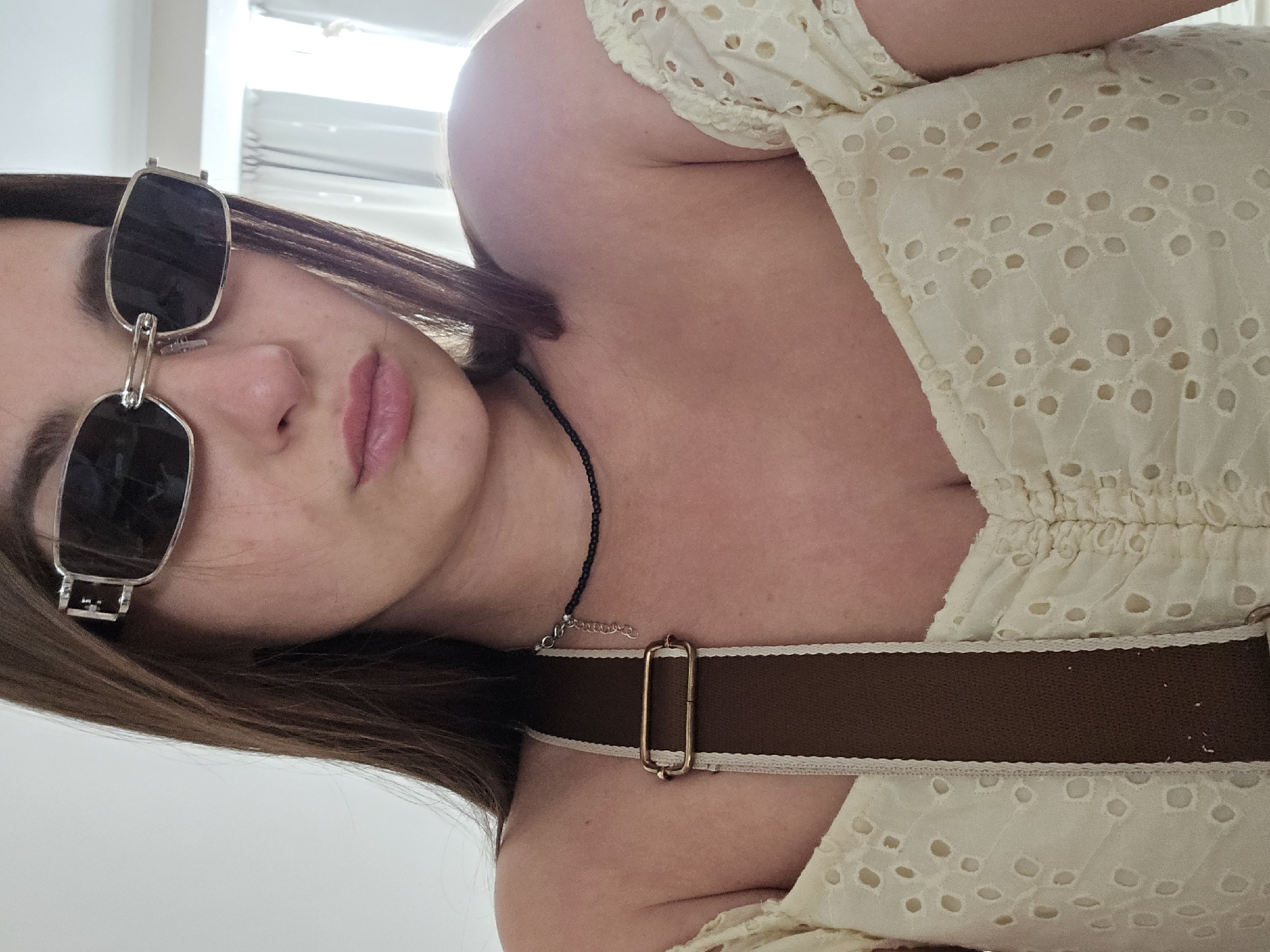A close-up, low-angle, portrait-style photograph shows a young woman from the chest up. She is facing towards the viewer's left, with her head tilted back and her eyes covered by black, rectangular sunglasses with silver frames. Her lips are parted slightly and appear to have a light pink lipstick on them. Her brown hair is visible around her face and shoulder.
She is wearing a light yellow, eyelet lace top with a ruffled neckline and off-the-shoulder sleeves. A dark brown strap with a cream-colored trim runs diagonally across her chest, secured by a rectangular gold buckle. A thin black beaded necklace with a small silver pendant is also visible around her neck.
The lighting suggests daytime, possibly indoors near a window with diffused light, indicated by the bright background and subtle shadows. The background is a plain, light-colored wall, possibly a room in a home. The overall mood is casual and relaxed. There is no visible text in the image. The provided location context of Petropavlivska Borshchahivka, Ukraine, cannot be determined from the image itself.