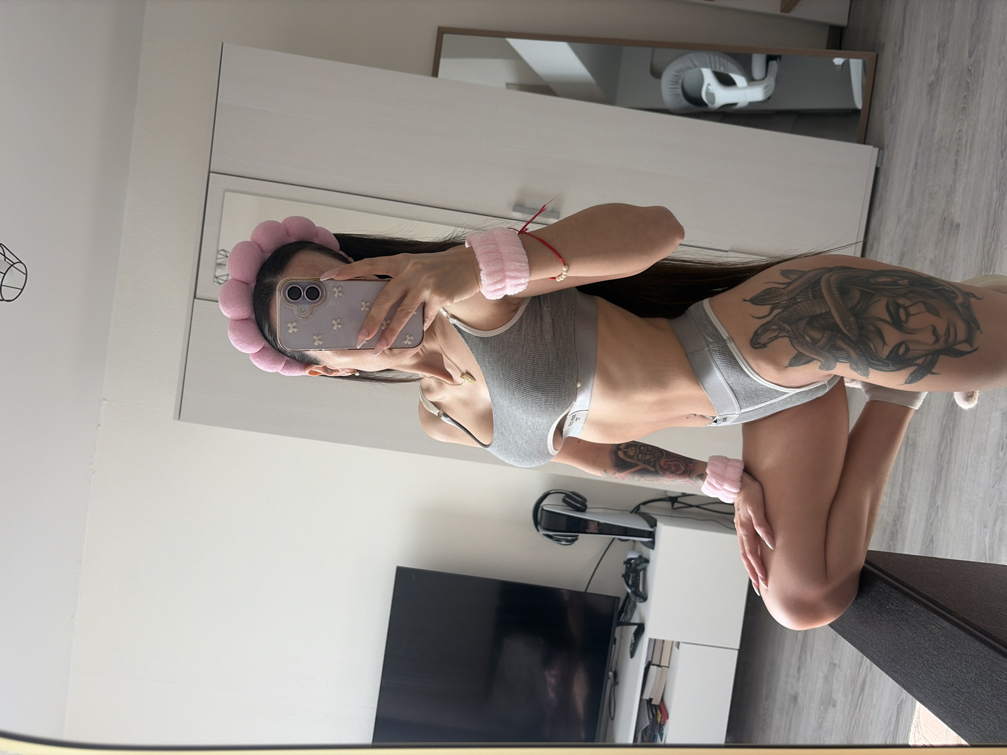 A young woman is captured in a mirror selfie, posing indoors in what appears to be a modern residential setting, likely in Saint Petersburg, Russia. She holds a light-colored smartphone with a floral-patterned case up to the mirror, obscuring part of her face. Her long, dark hair falls over her shoulders. She wears a pink, puffy headband and matching wristbands, along with a grey ribbed crop top and matching high-waisted underwear. A delicate gold necklace is visible around her neck, and she also has a red string bracelet with a pearl on her right wrist. Her long, light-colored manicured nails are prominent as she holds the phone.
Notable details include a large, intricate tattoo featuring what appears to be snakes or a Medusa-like figure on her right side/hip area, and another tattoo is partially visible on her left arm. The room's interior features light-colored walls and a wardrobe or cabinet. In the background reflection, a baby car seat or carrier can be seen on a shelf. A PlayStation 5 console and headphones are visible on a white stand, next to a large, turned-off television screen. The flooring is a light-colored wood or laminate. The overall scene suggests a casual, private moment in a well-kept home. No specific text is clearly visible on any objects or clothing. The lighting suggests daytime.