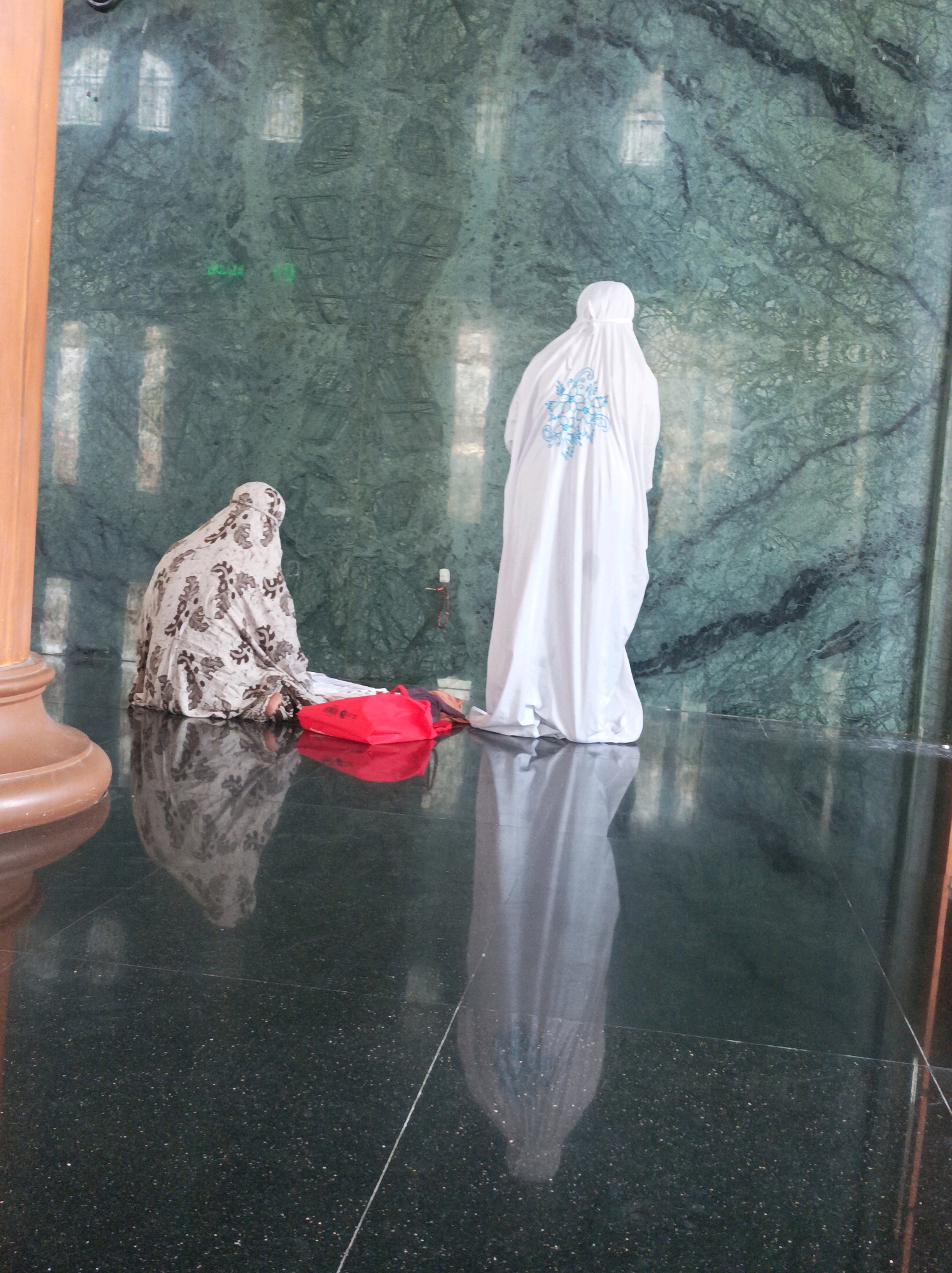 Two individuals, dressed in traditional Islamic attire, are engaged in prayer within a building featuring green marble walls and a dark, reflective floor. The person on the left is wearing a patterned abaya and is seated, with a red bag beside them. The person on the right is wearing a white abaya with light blue embroidery on the back and is also in prayer. Their reflections are visible on the polished floor. The environment appears to be a place of worship, likely a mosque, given the context and attire. The location is specified as Rengel, Indonesia. The lighting suggests an indoor setting, possibly with natural light filtering in from unseen windows, as evidenced by the reflections on the wall.