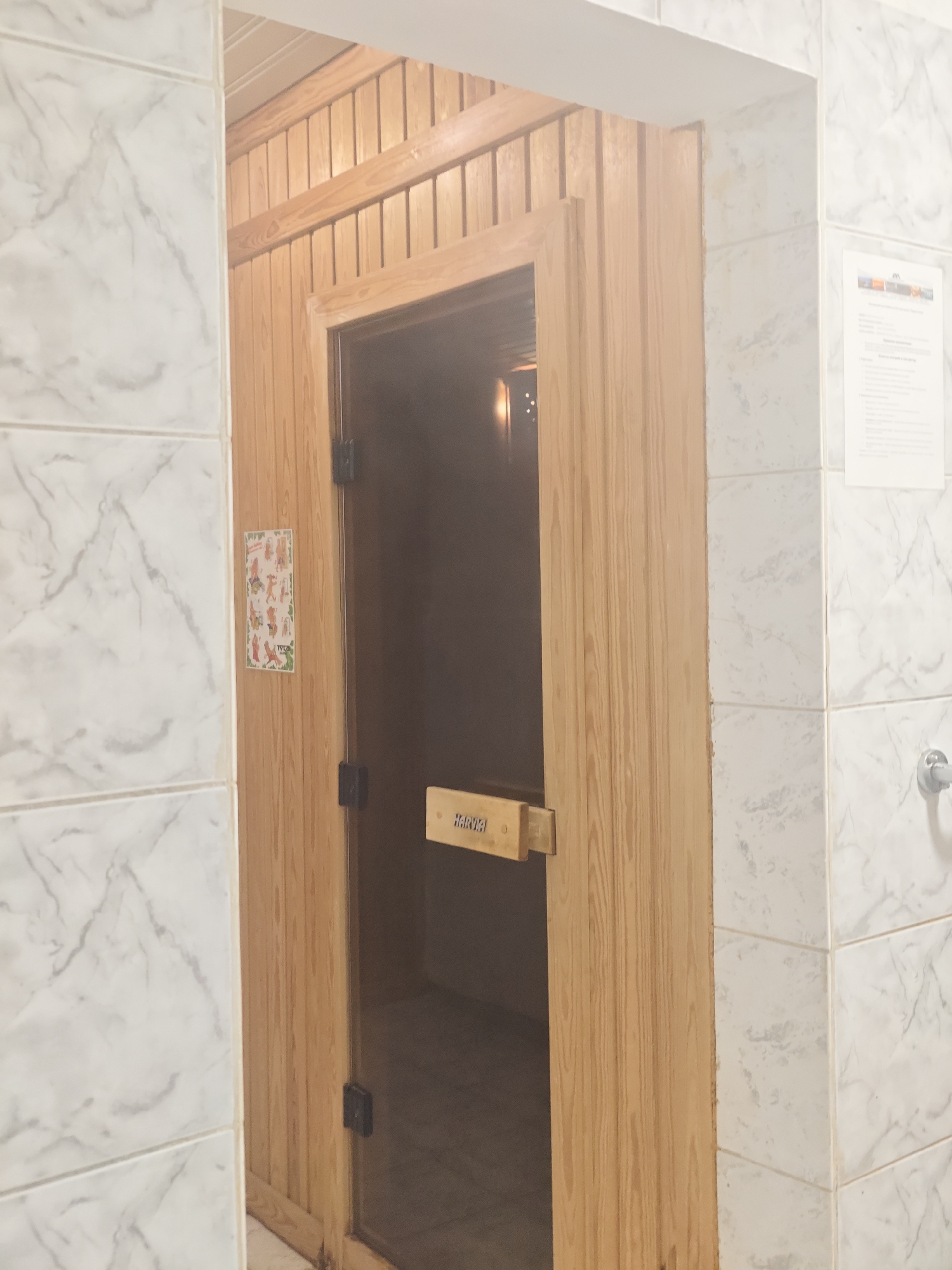 Indoor setting in Kozloduy, Bulgaria. A wooden sauna door is ajar. The adjacent walls are tiled with white marble-patterned tiles. A sign with human figures is affixed to the door frame. A Harvia brand handle is present. A safety information sheet is attached to the tiled wall. A silver tap is visible on the right tiled wall.