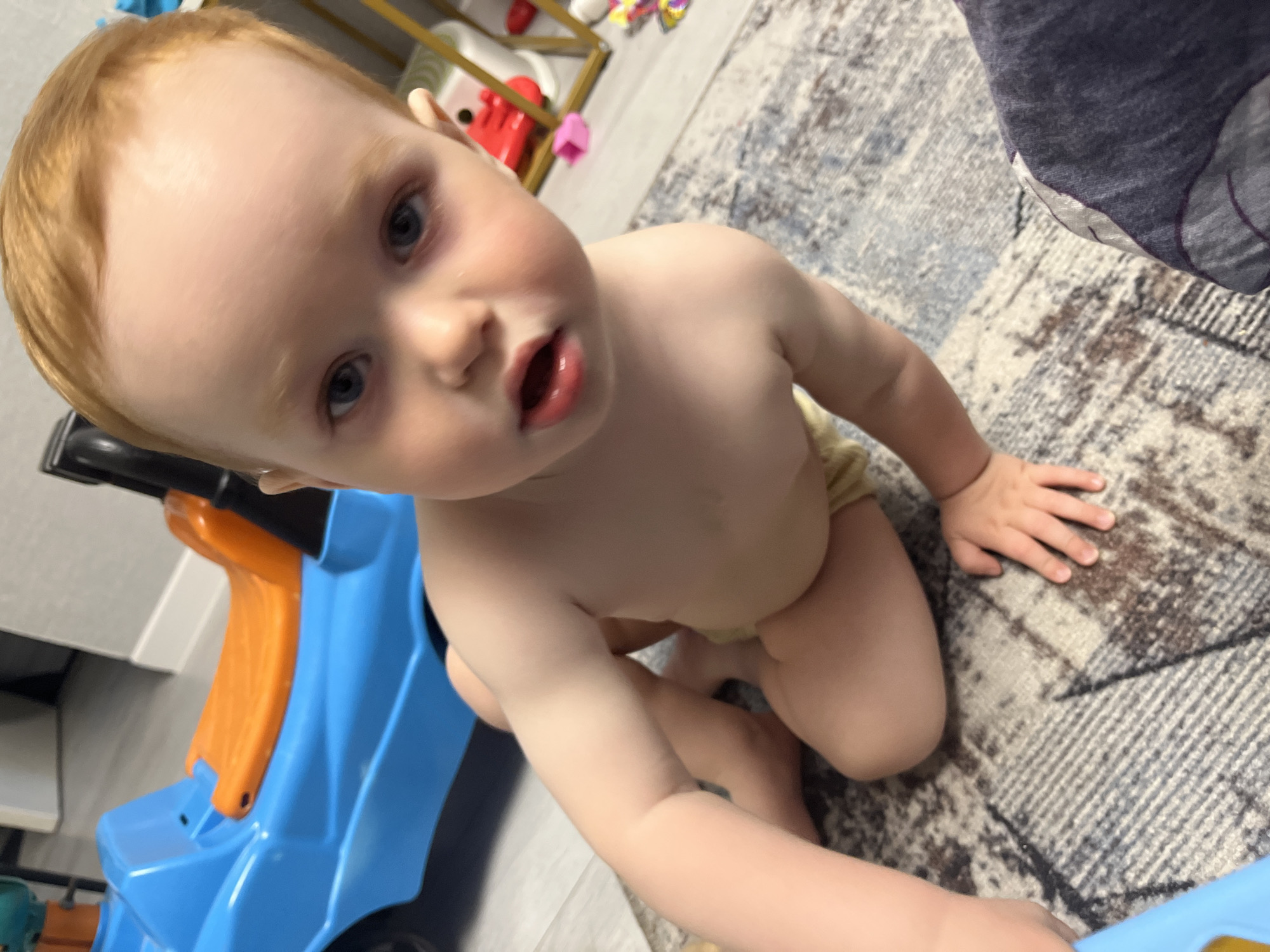 A baby with red hair and blue eyes is kneeling on a rug with a blue and orange ride-on toy. The location is potentially in Chaikovsky, Russia, but there is nothing in the image to confirm this. The baby is looking up at the camera with its mouth slightly open. The rug has a brown and gray pattern. Part of a metal shelf with toys is visible in the background. A gray and blue cushion is partially visible in the upper right corner.