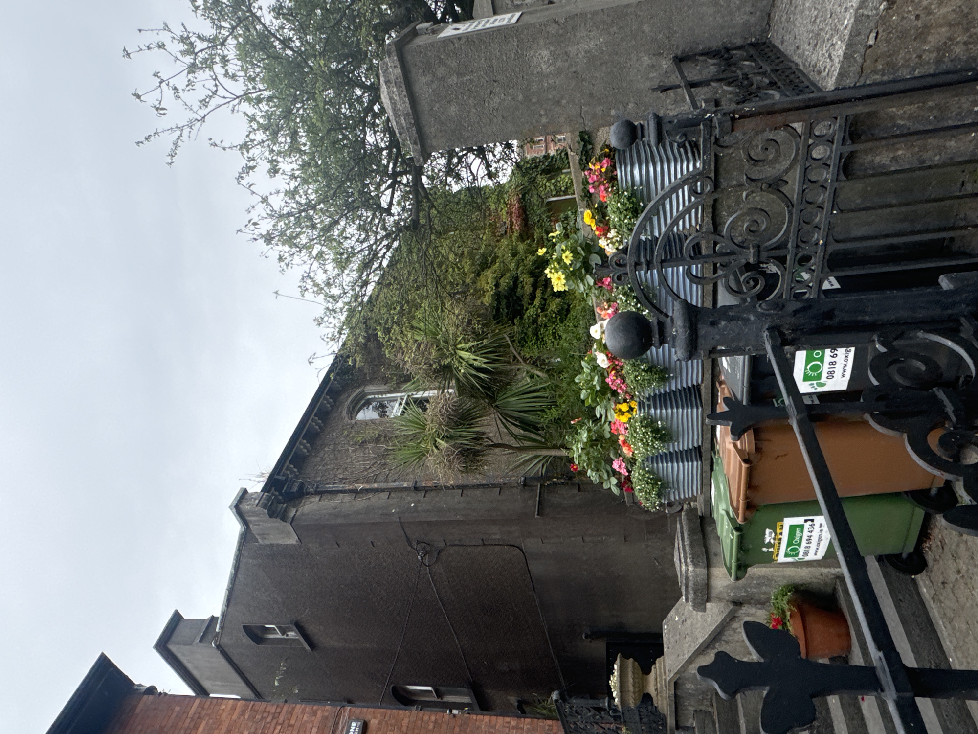 The image depicts an outdoor scene with a focus on a row of modern, metallic planters filled with colorful flowers. These planters are positioned on a raised area, possibly a wall or a terrace, behind a decorative black metal gate. To the left of the planters, a set of stone steps leads up to a dark-colored building with a chimney. Below the planters and in front of the gate, several wheelie bins are visible, including a green one and a brown one, both bearing stickers with a phone number and website address. A small terracotta pot with a red flower is on the steps to the left. The background reveals more of the building, dense greenery, and the sky, which is overcast. The scene appears to be in an urban setting, likely a residential area.
The original image was captured in Waterford city, Ireland.
