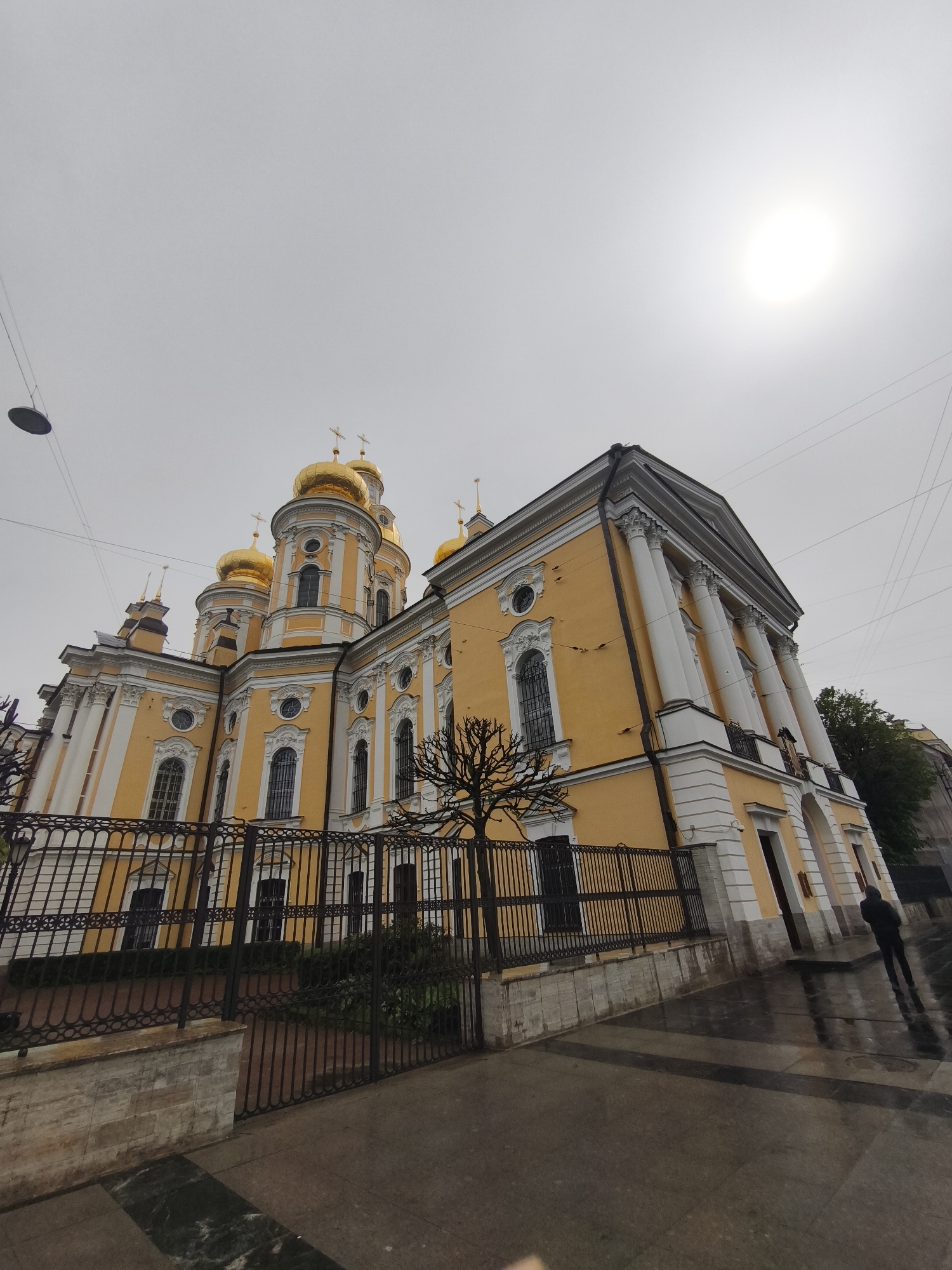 The image depicts a yellow and white building with golden domes, suggesting a church or cathedral. A black metal fence surrounds the property. The ground appears wet, possibly from rain. In the foreground, a person in dark clothing stands near the building. The sky is overcast with the sun visible as a bright, diffused circle. Overhead wires are strung above the building. The location is in an unknown city, as no identifying details are present.