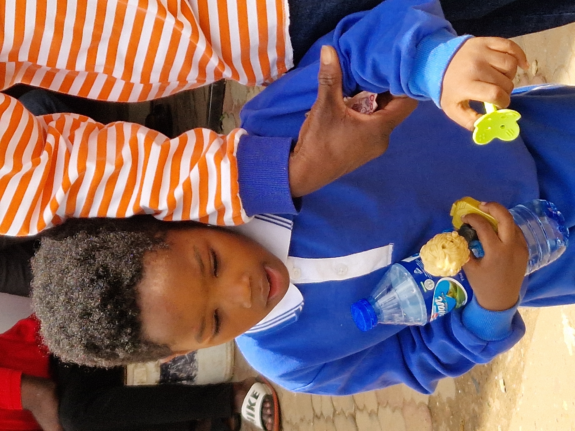 A young Black child wearing a blue collared shirt with white trim is holding a plastic water bottle and a small yellow toy. The child's hair is dark and curly, and their eyes are looking down. Another person, partially visible in the background, is wearing an orange and white striped shirt. They are outdoors on a paved surface, possibly a street or walkway. The lighting suggests it is daytime.